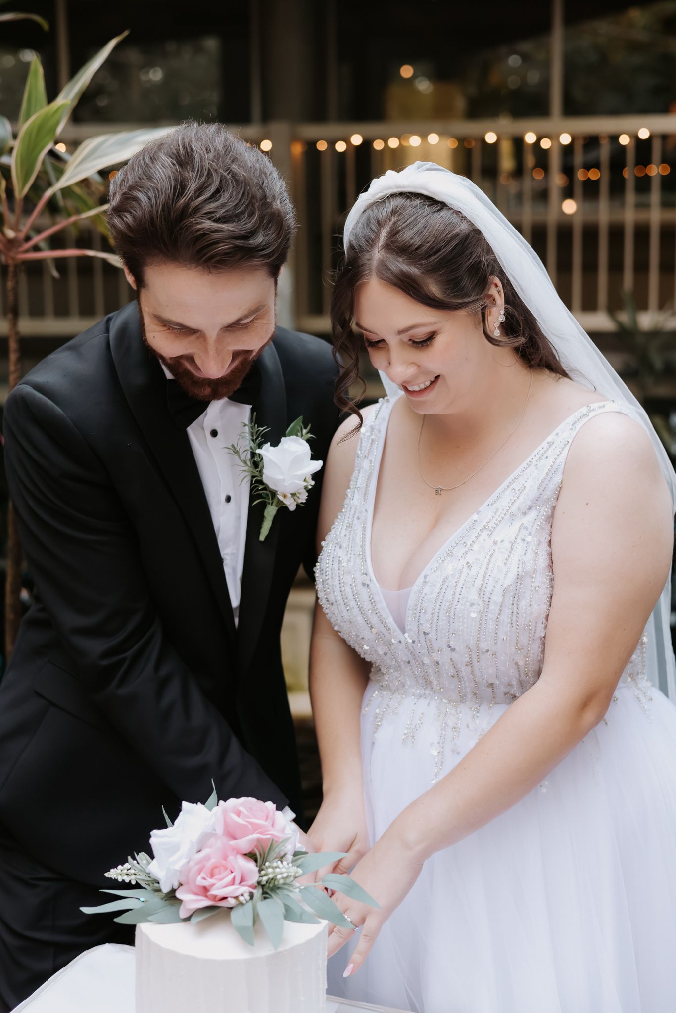 Bride and groom cutting their wedding cake adorned with pink roses, dressed in elegant wedding attire.