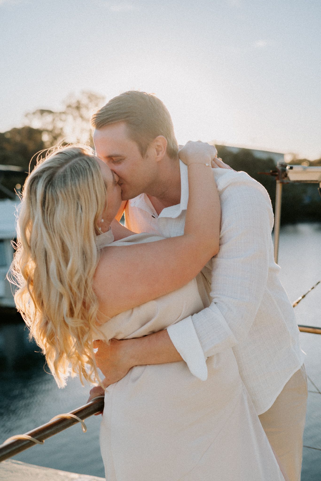 Couple sharing a romantic kiss at golden hour on a waterfront dock, dressed in elegant white outfits.