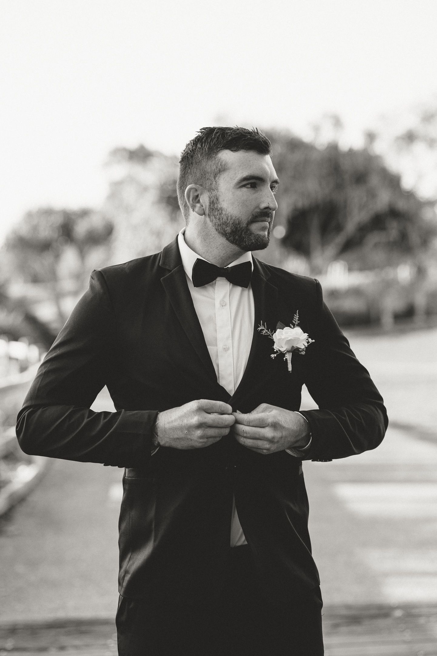 Groom in black tuxedo with bow tie and boutonniere buttoning jacket outdoors in black and white photo.