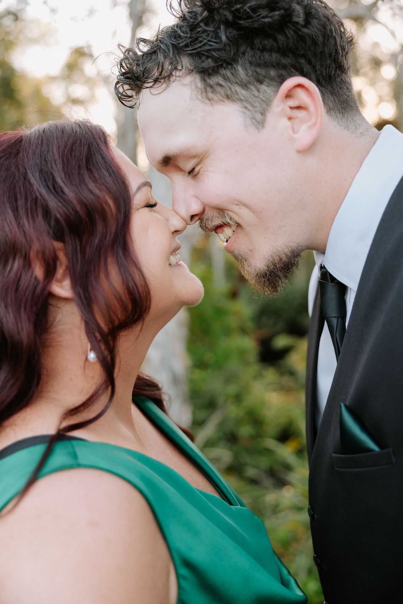 A smiling couple in formal attire share a romantic nose-to-nose moment outdoors, woman in green dress, man in black suit.
