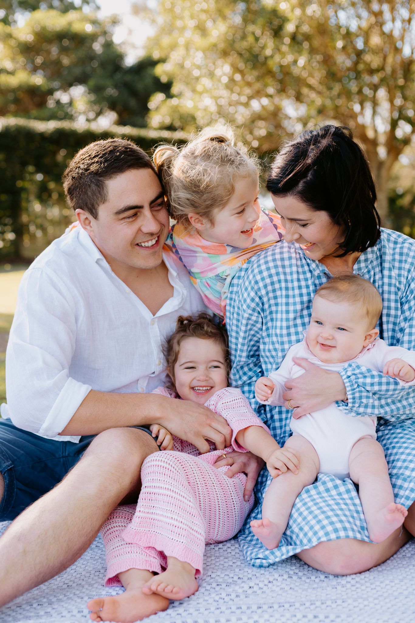 Happy family of five sitting outdoors on a blanket, parents smiling with three young children in a sunny garden.