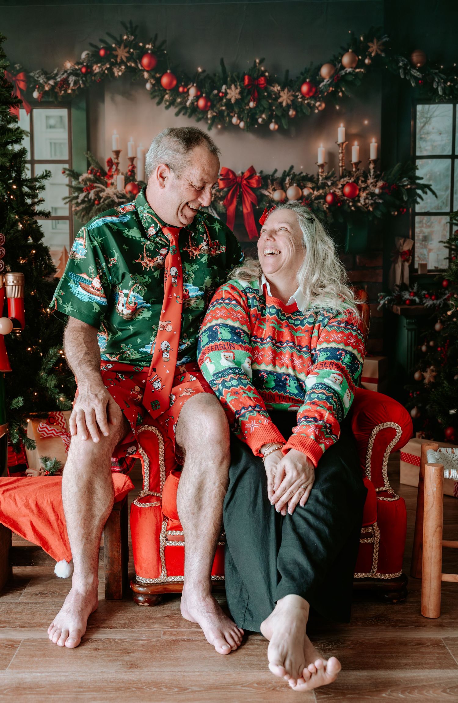Elderly couple laughing together in matching Christmas outfits on a red chair with festive holiday decorations.