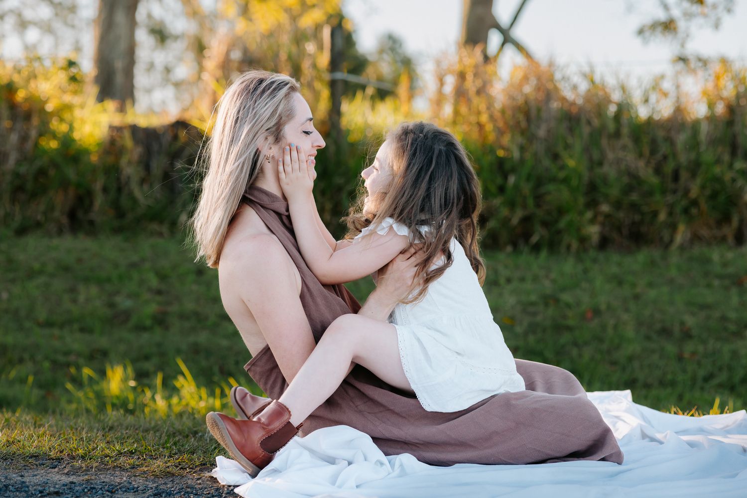 Two girls sitting on a white blanket outdoors, sharing a tender moment while holding each other's faces.