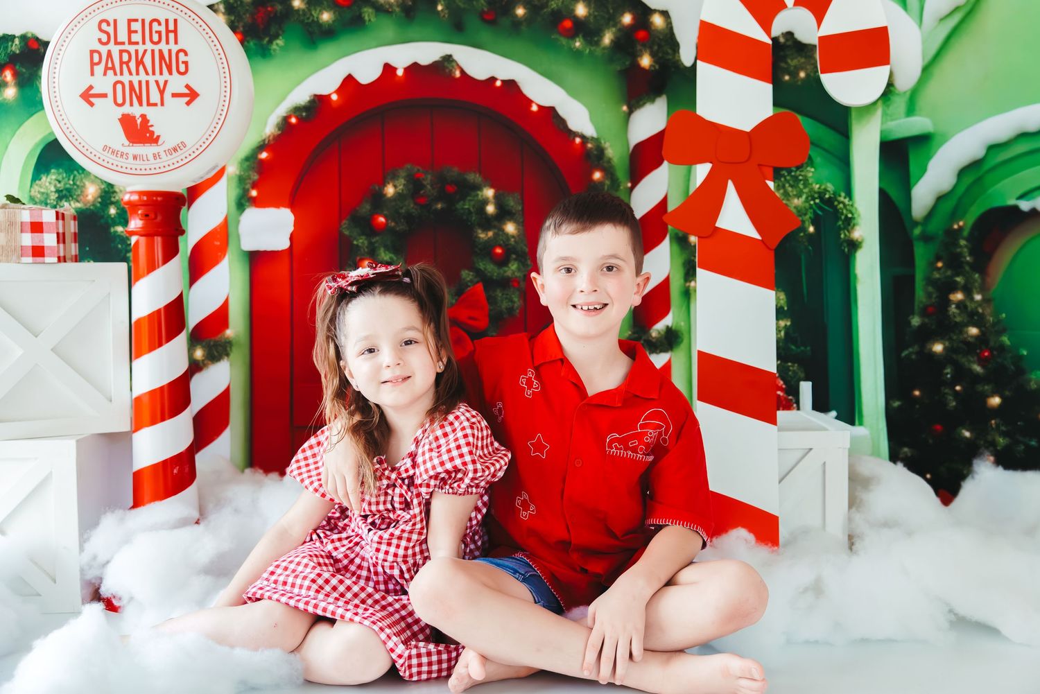 Two children in red holiday outfits pose before a festive Christmas candy cane backdrop with a Sleigh Parking Only sign.