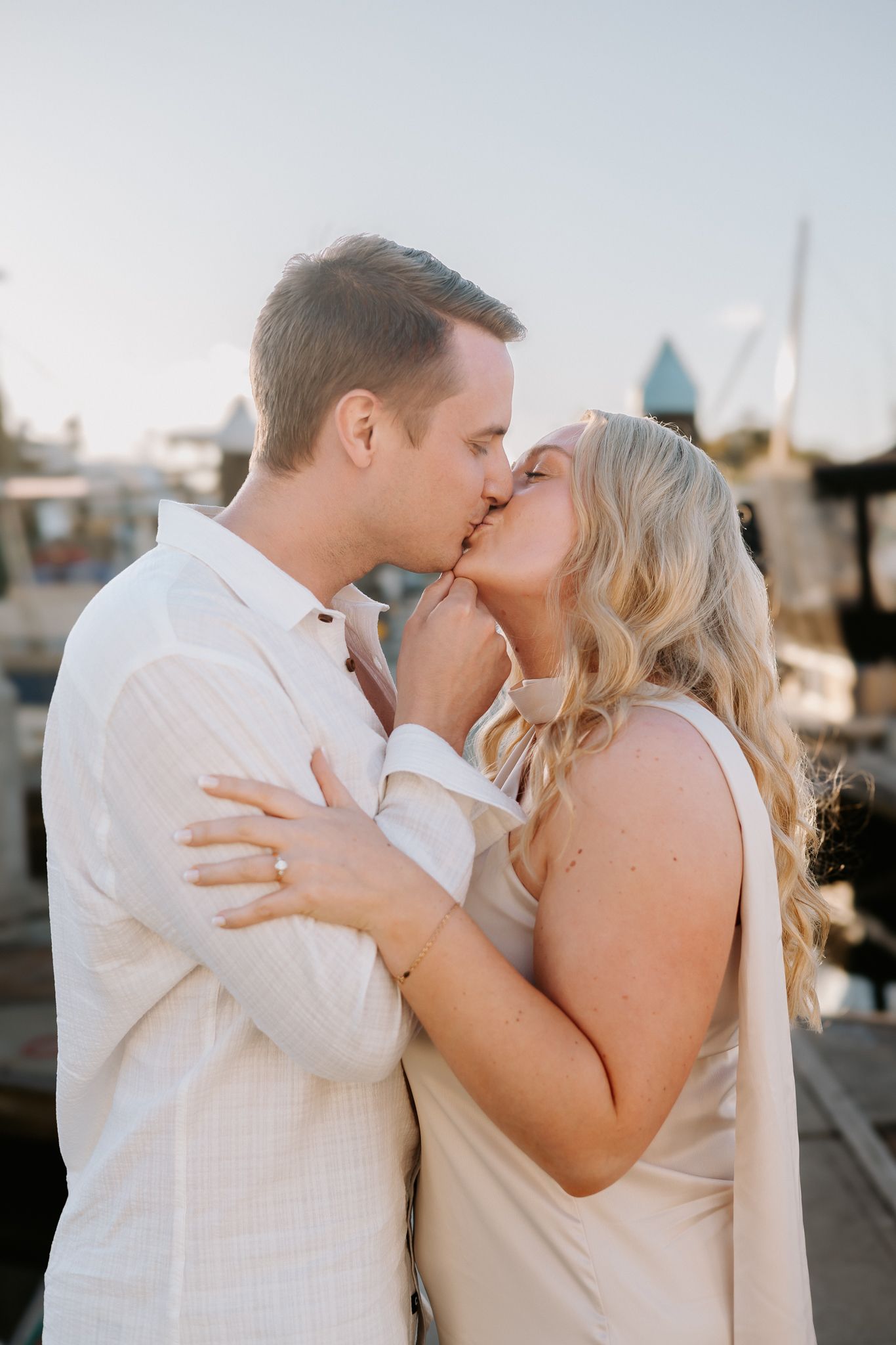 Couple sharing a romantic kiss at a marina during golden hour, dressed in elegant white outfits.