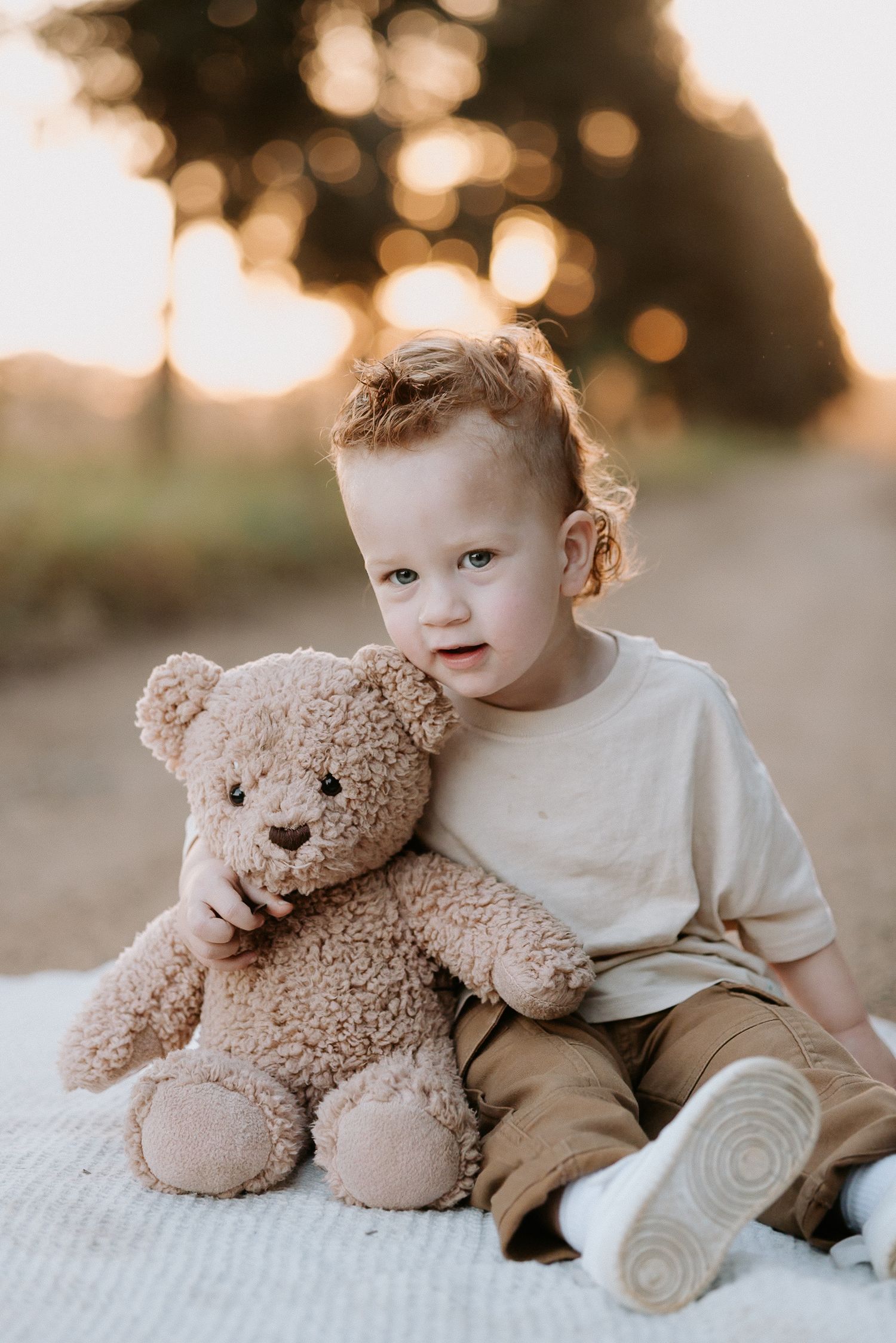 Toddler boy with red hair hugging a fluffy teddy bear outdoors at golden hour sunset.