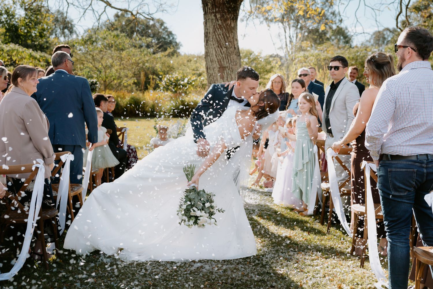 Bride and groom kissing while guests toss confetti at an outdoor wedding ceremony under a large tree.