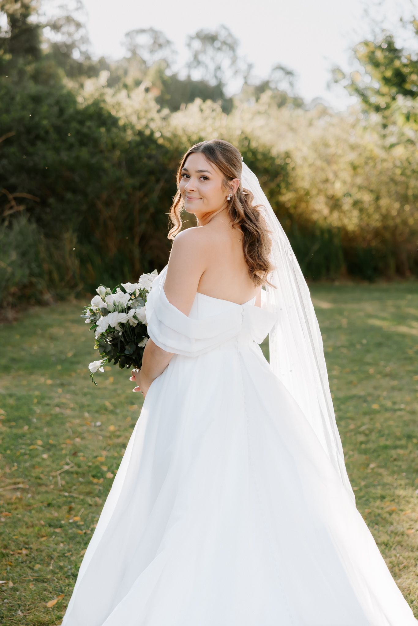 Bride in white off-shoulder gown with veil holds floral bouquet, smiling over shoulder in lush garden setting.