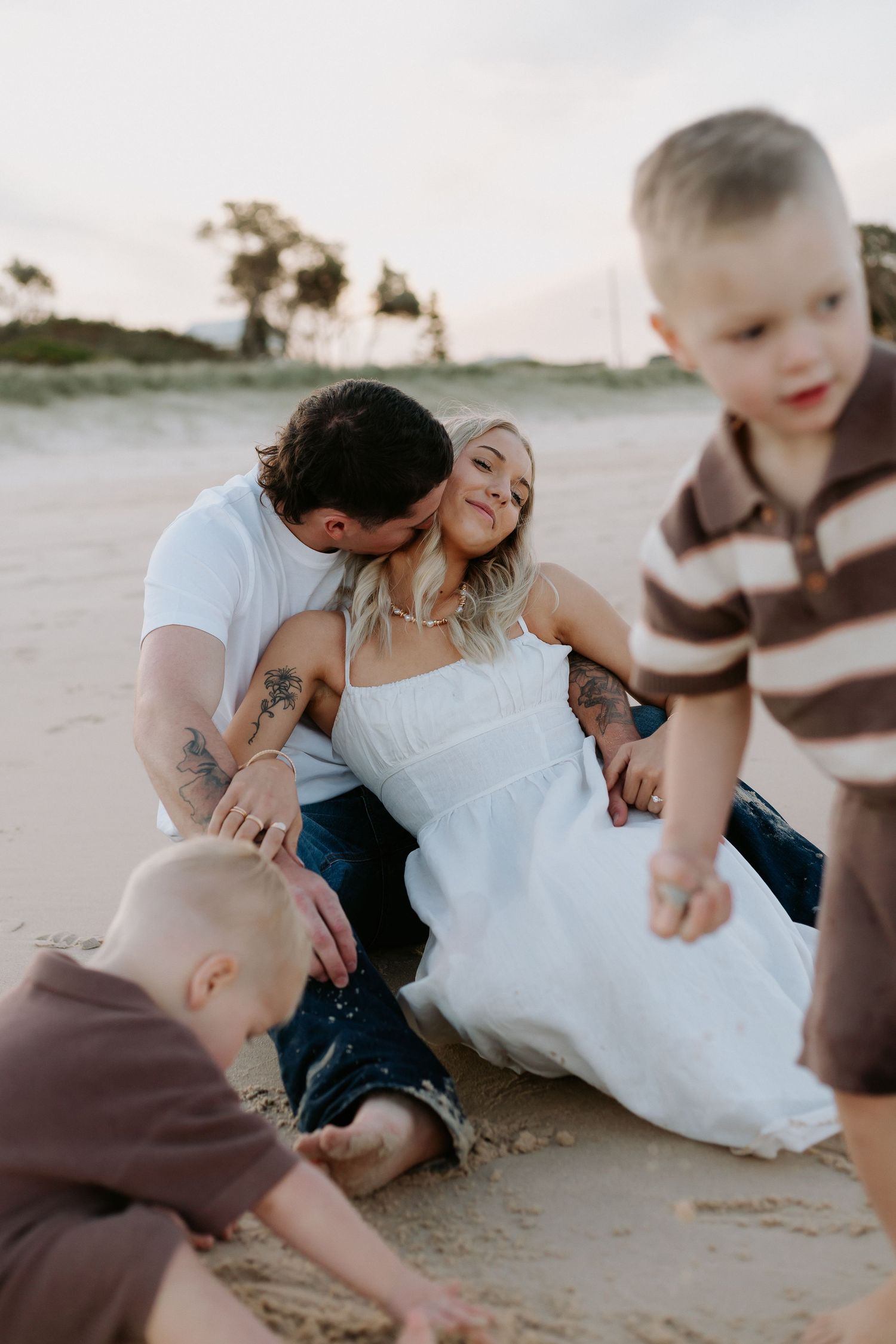 A couple sits on a sandy beach with two young boys playing around them at sunset.