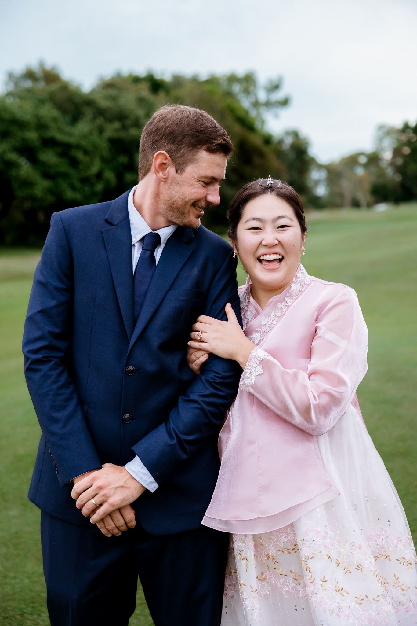 Smiling couple in wedding attire, man in navy suit and woman in pink Korean hanbok, embracing outdoors on green lawn.