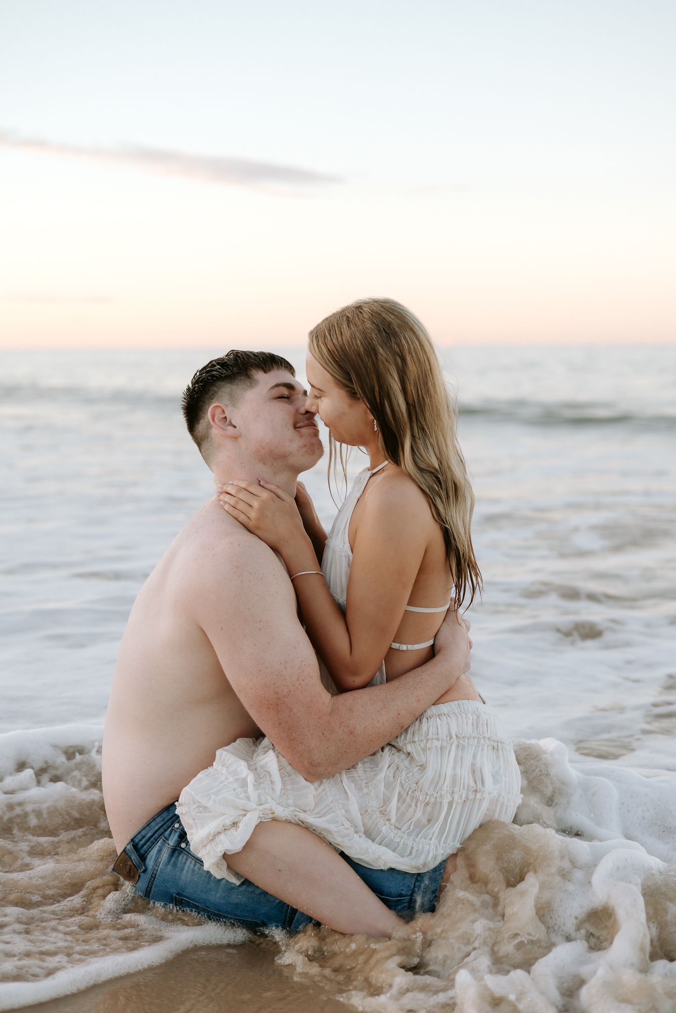 Romantic couple embracing in ocean waves at sunset on a sandy beach, woman in white dress and man in jeans.
