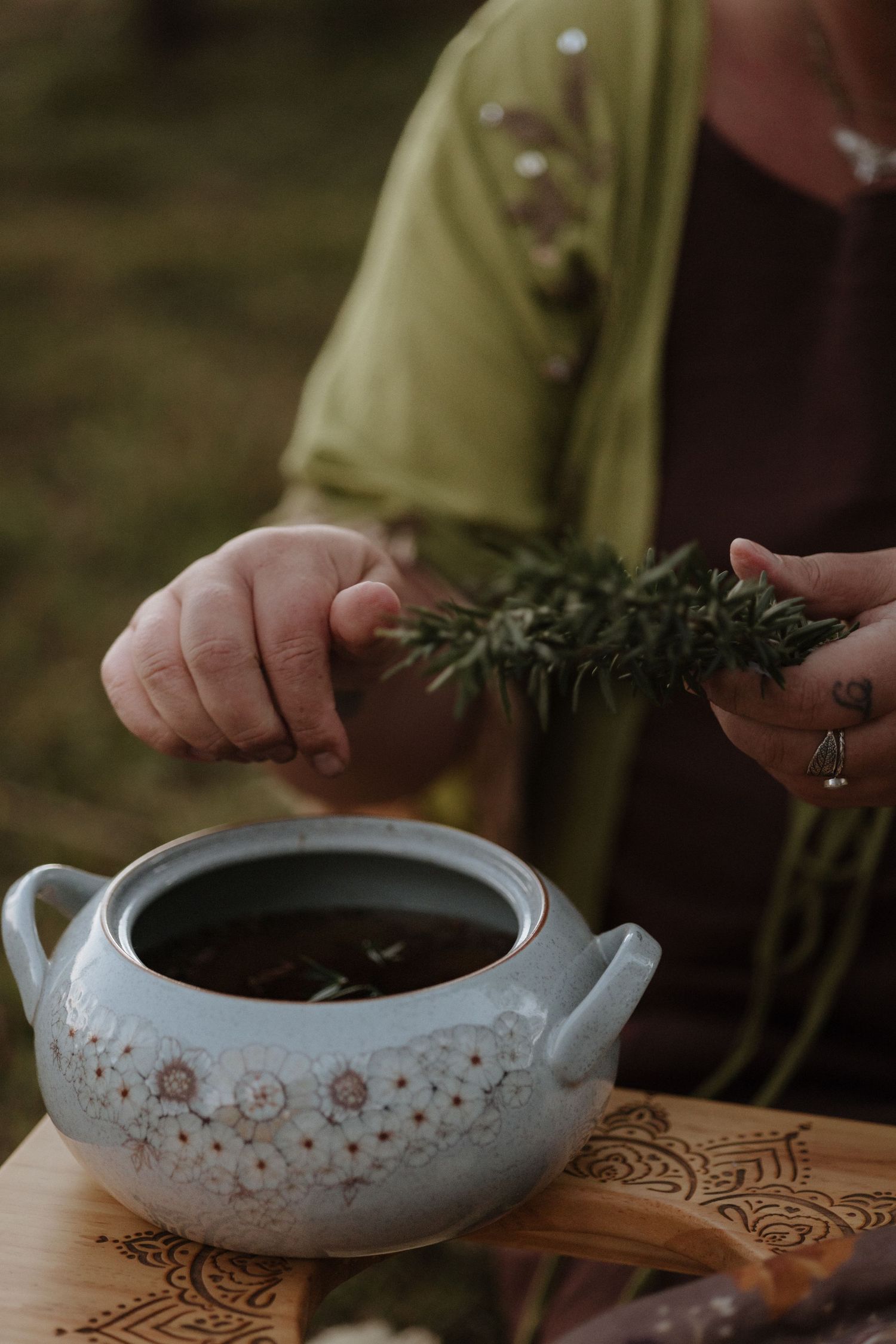 Woman adding fresh rosemary sprigs to a decorative ceramic pot on a wooden board outdoors.