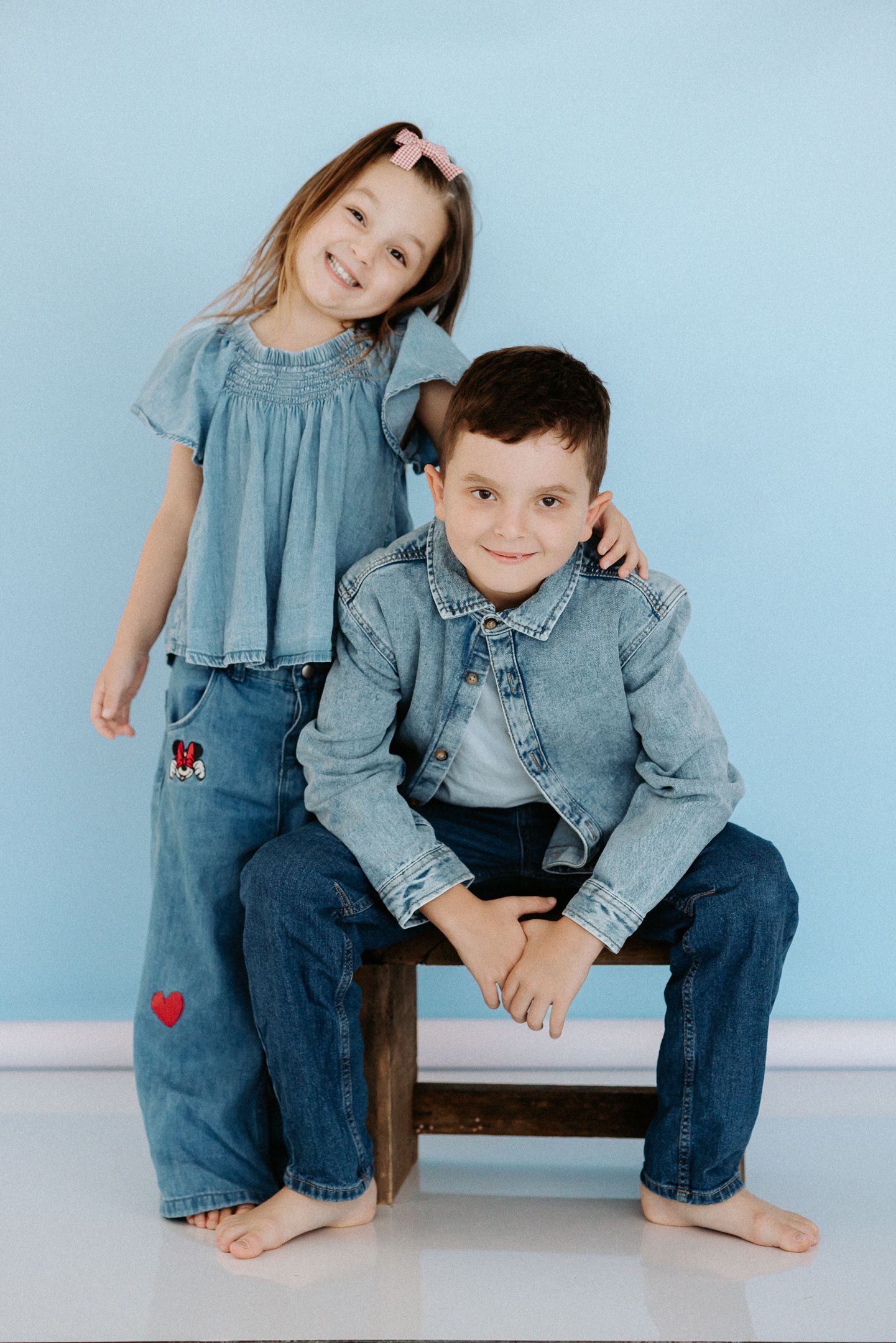 Two children in denim outfits pose barefoot against a light blue wall with one sitting on a wooden stool.