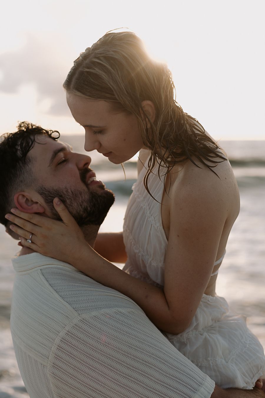 Man lifting woman in white dress on beach at sunset, sharing a romantic almost-kiss by ocean waves.