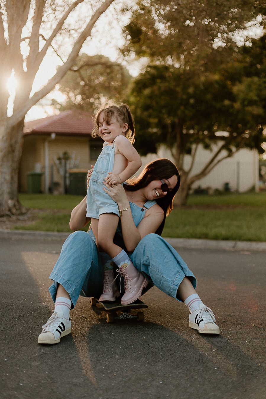 Parent and child sitting on skateboard together at sunset in matching blue outfits and white sneakers.