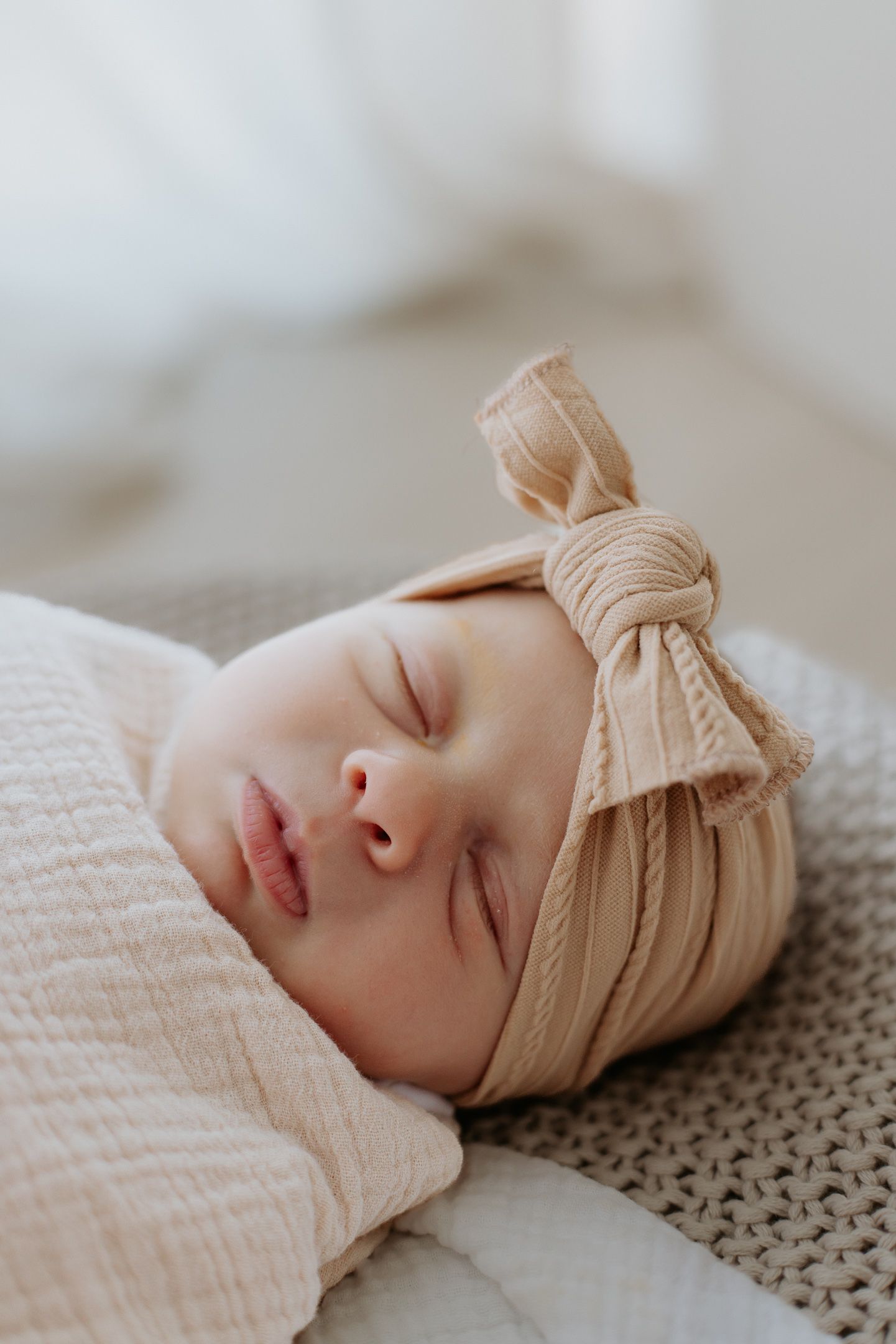 Sleeping infant wearing soft beige bow headband rests peacefully on textured blanket in gentle natural lighting.