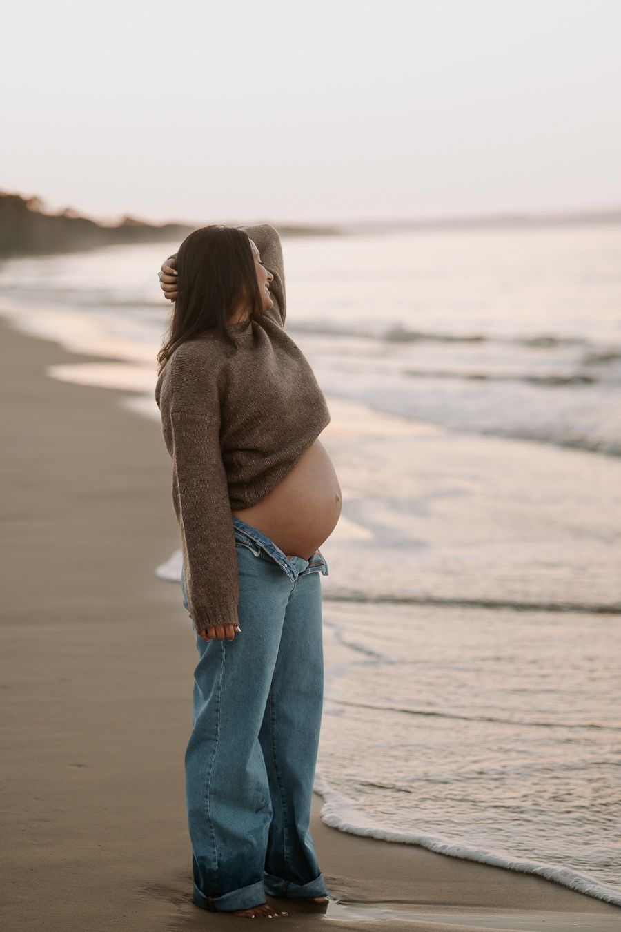 A pregnant person in a sweater and jeans stands on a misty beach at sunset, looking out at the ocean waves.
