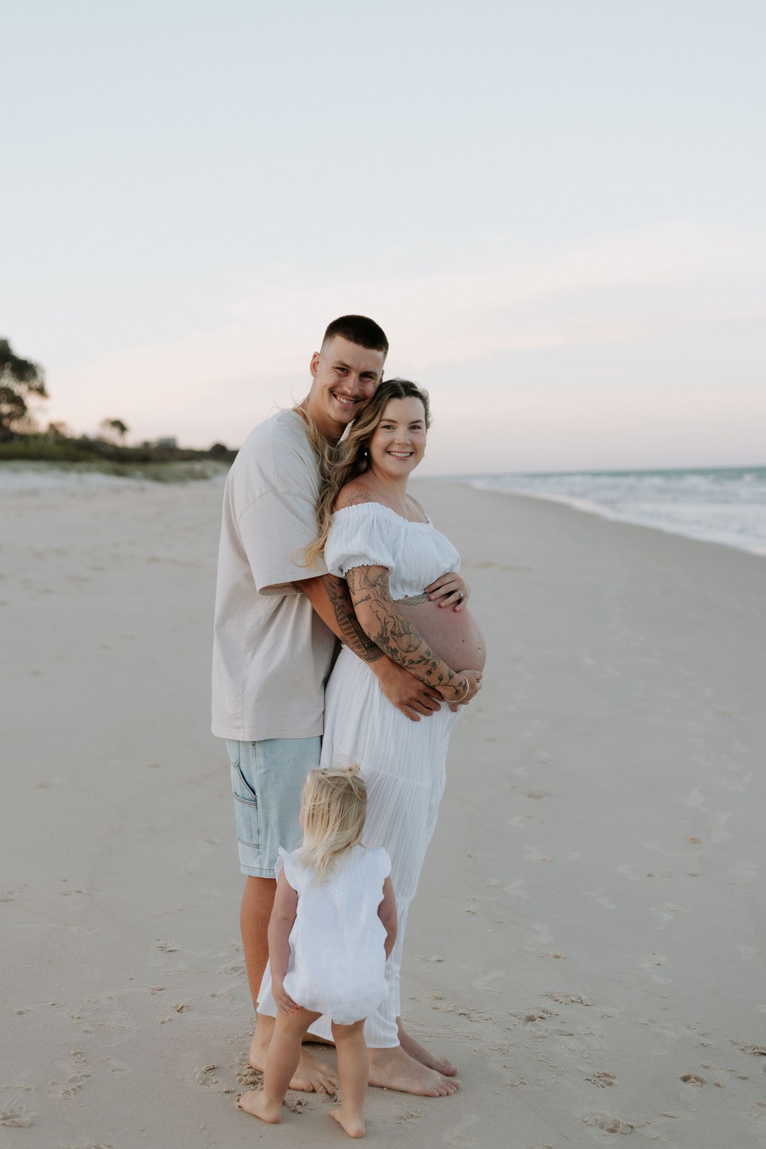 A couple embraces on a serene beach at sunset, with the woman wearing a flowing white dress showing her pregnant belly.