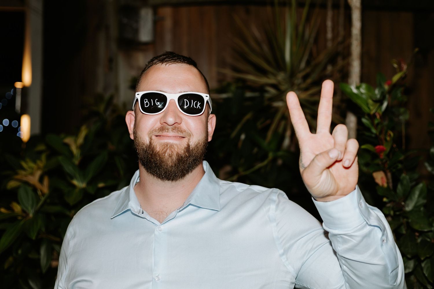 Guest in light blue shirt and sunglasses making peace sign at outdoor event.