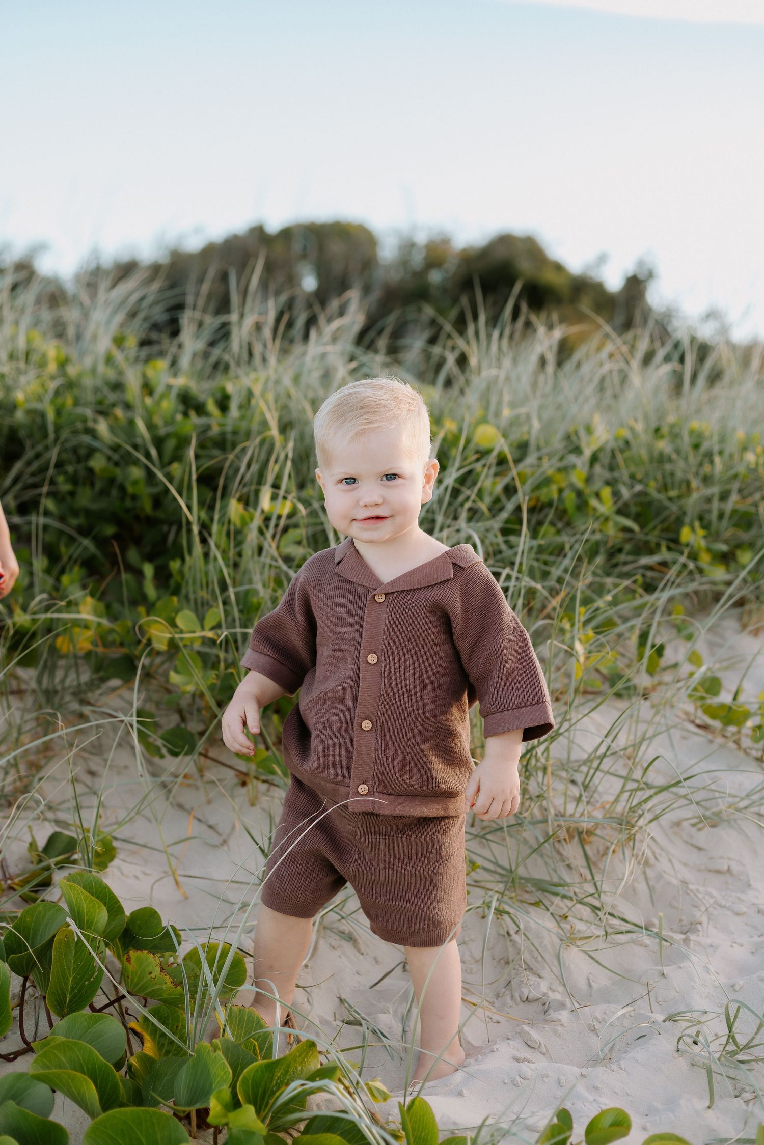 Toddler boy in brown shorts set standing on sandy beach dunes with green coastal plants.