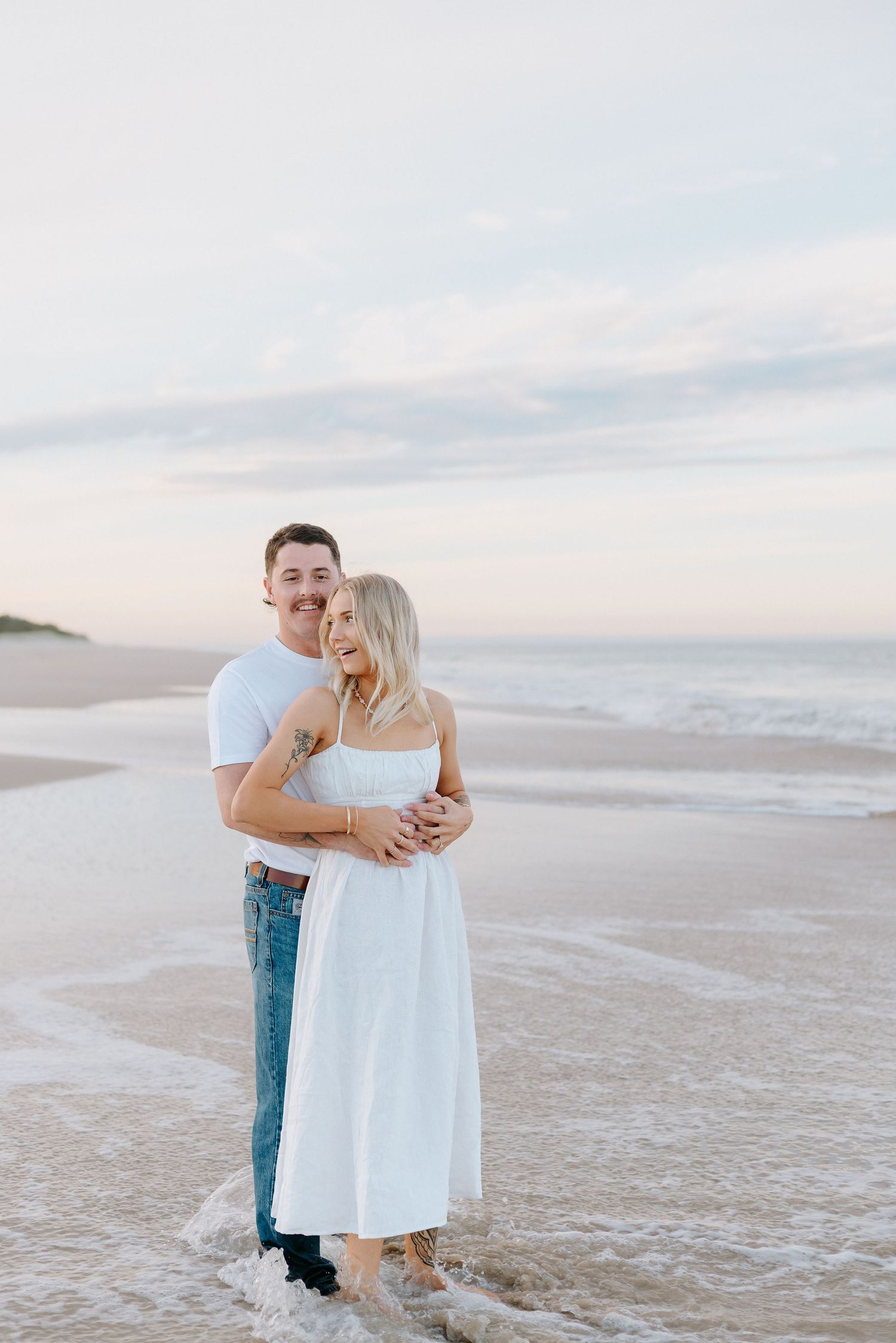 A couple embraces on a sandy beach at sunset, the woman in a white dress and the man in a white tee and jeans.