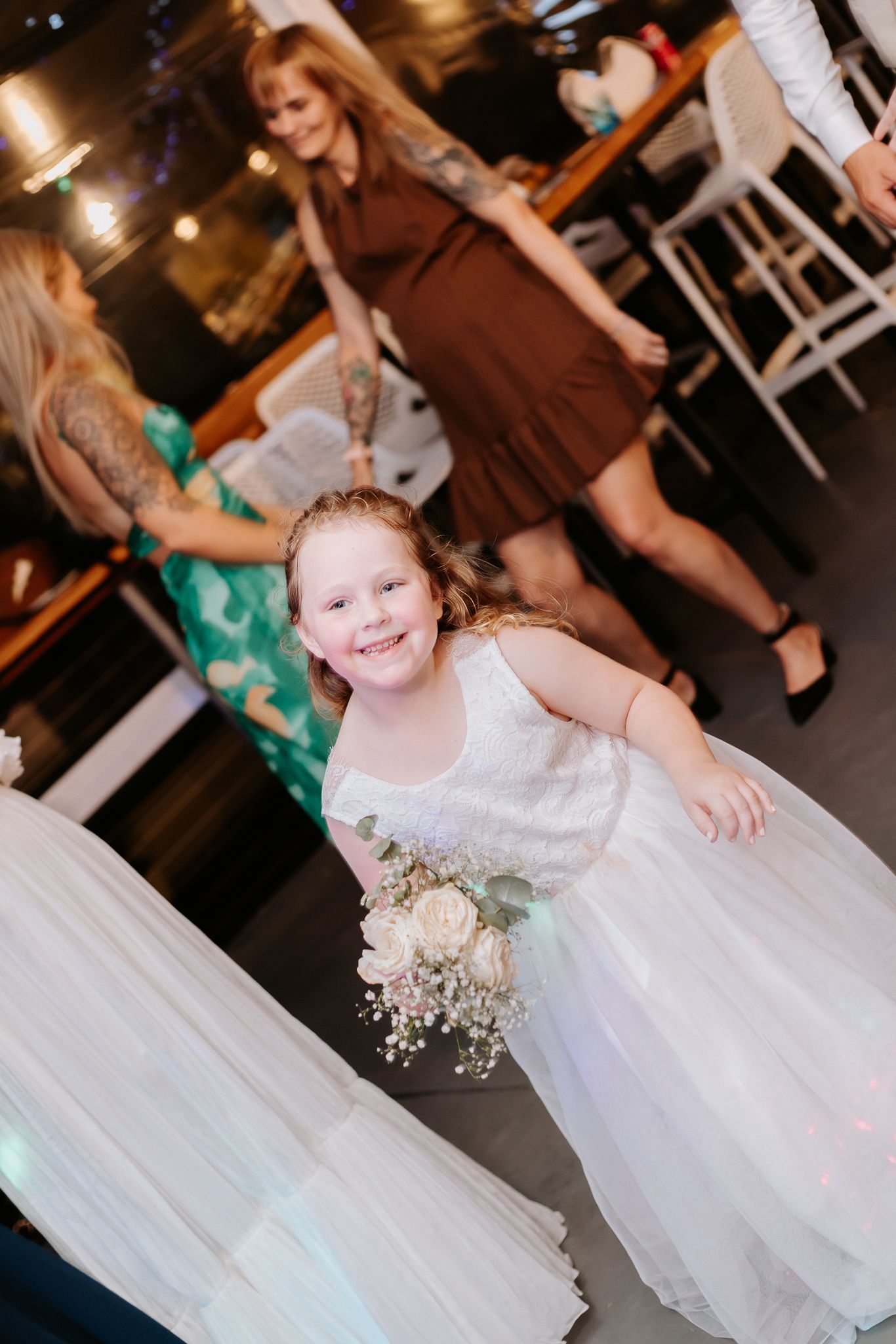 Smiling young flower girl in white dress holding bouquet at a wedding reception venue.