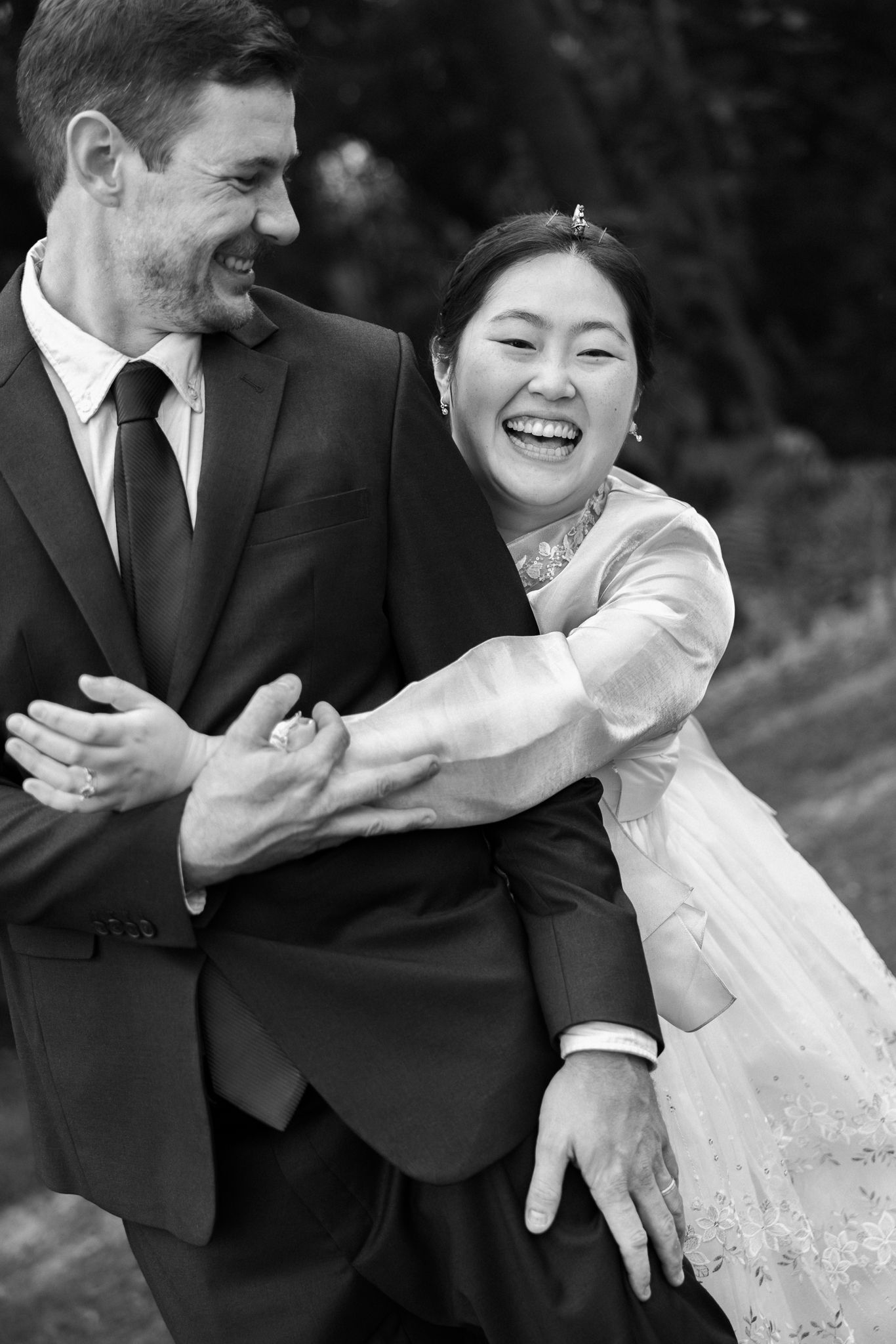 Black and white photo of a laughing bride in white dress embracing a smiling groom in dark suit outdoors.