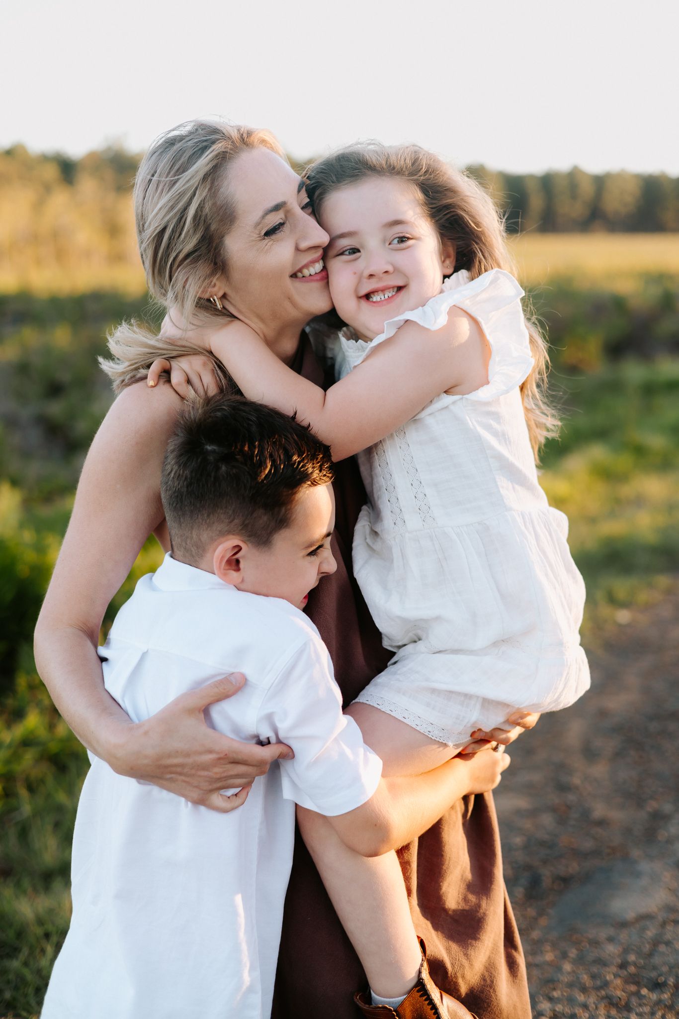 A smiling mother hugs her two young children outdoors in a sunlit field, sharing a warm family embrace.