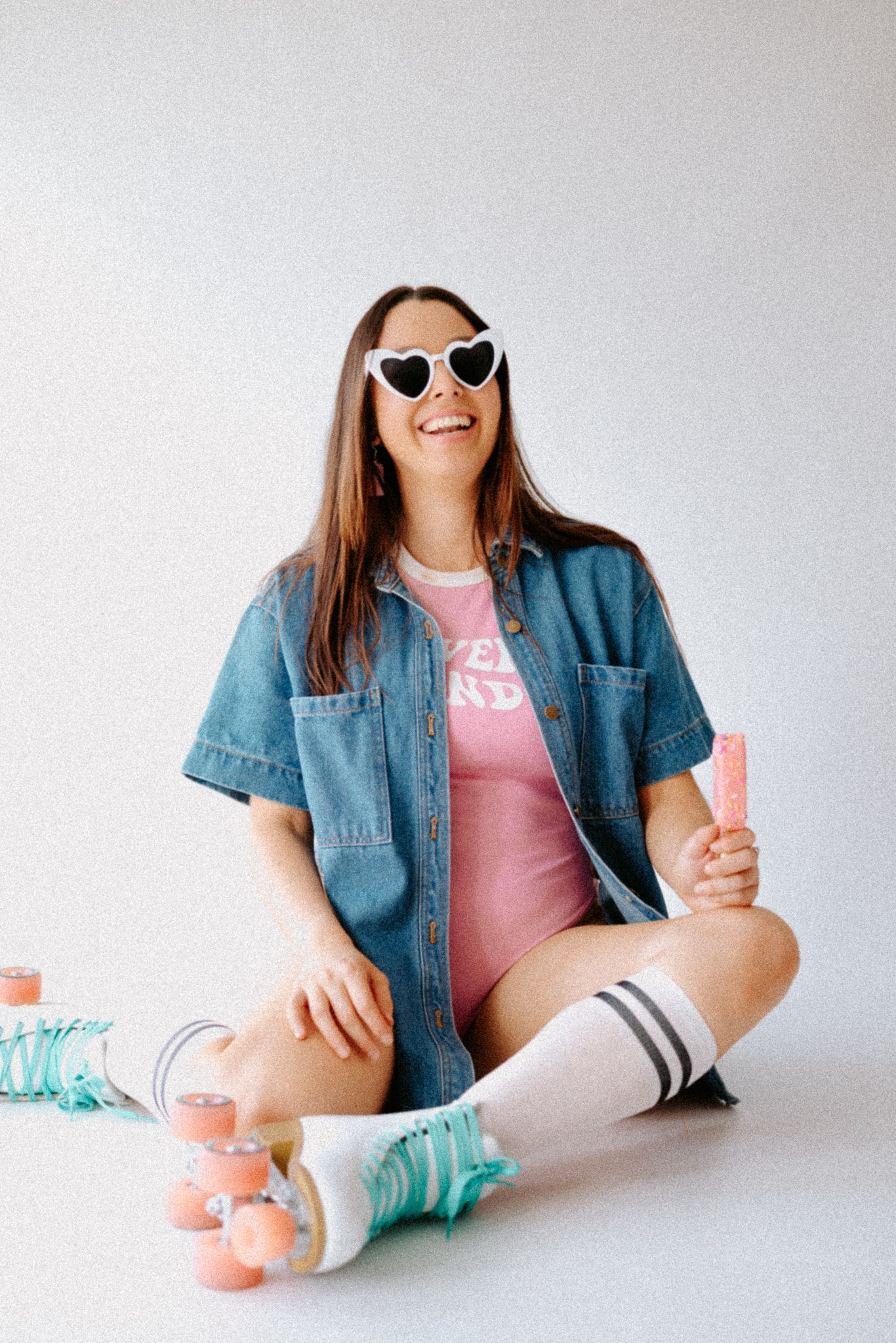 Smiling woman in denim shirt, pink romper, heart sunglasses, roller skates, holding a popsicle while sitting on white floor.