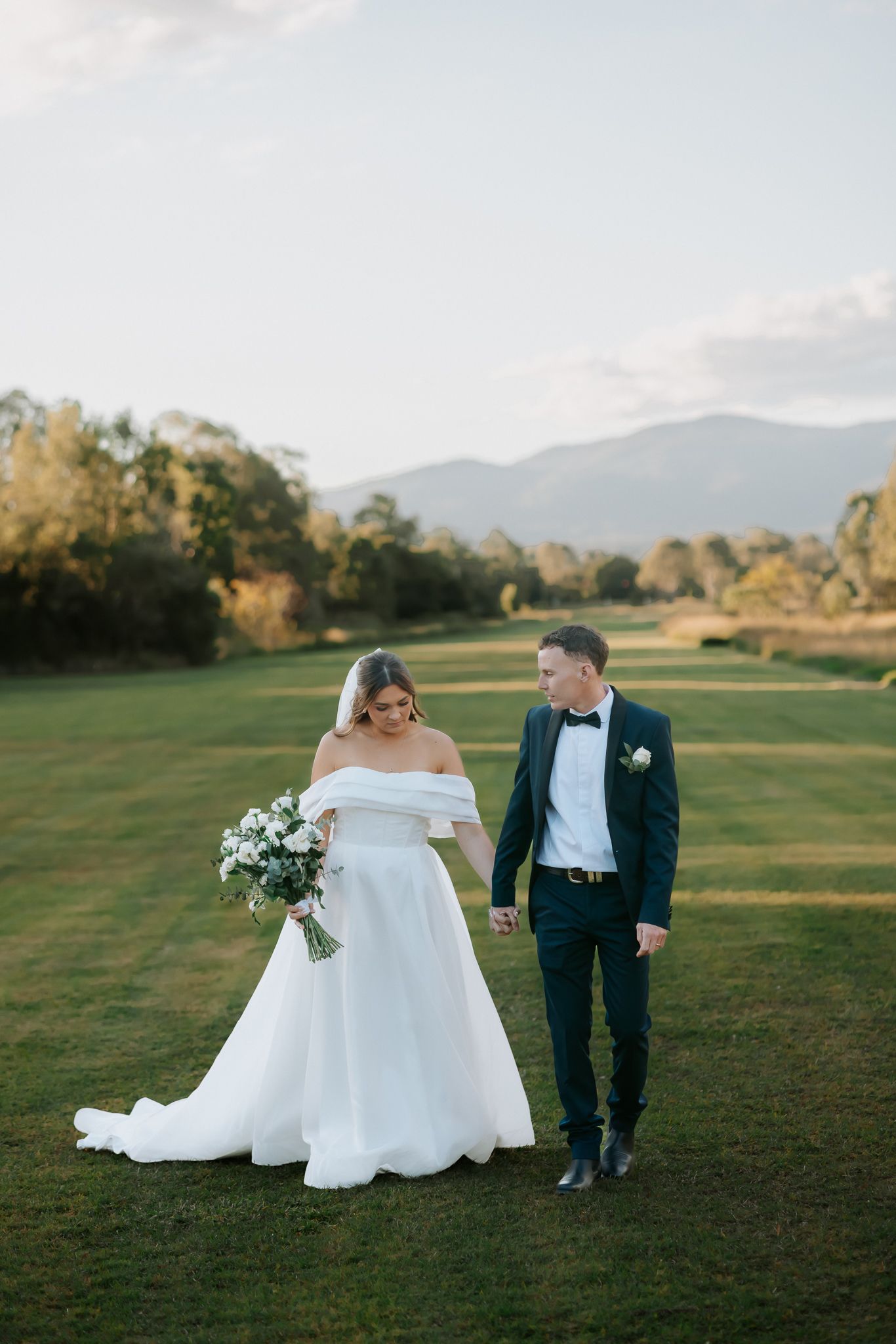 Bride and groom walking hand in hand on green lawn with mountain backdrop at golden hour wedding.