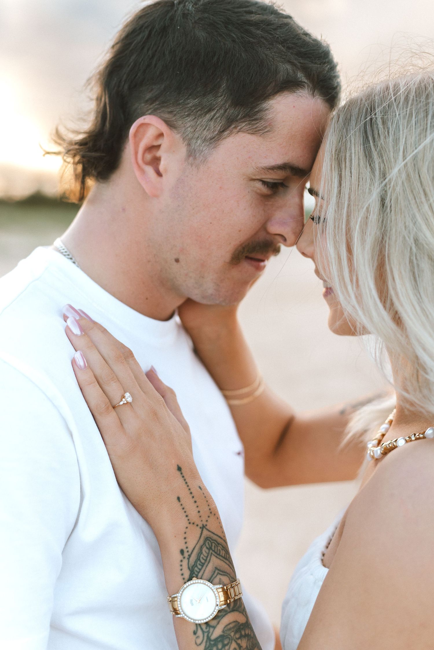 Romantic couple in white outfits touching foreheads at sunset, woman showing engagement ring and tattoo.