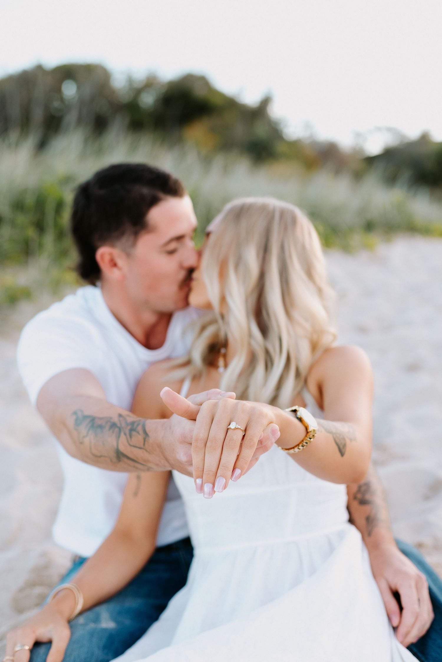 Couple kissing on beach while showing off engagement ring, woman in white dress with tattoos, sandy dunes background.