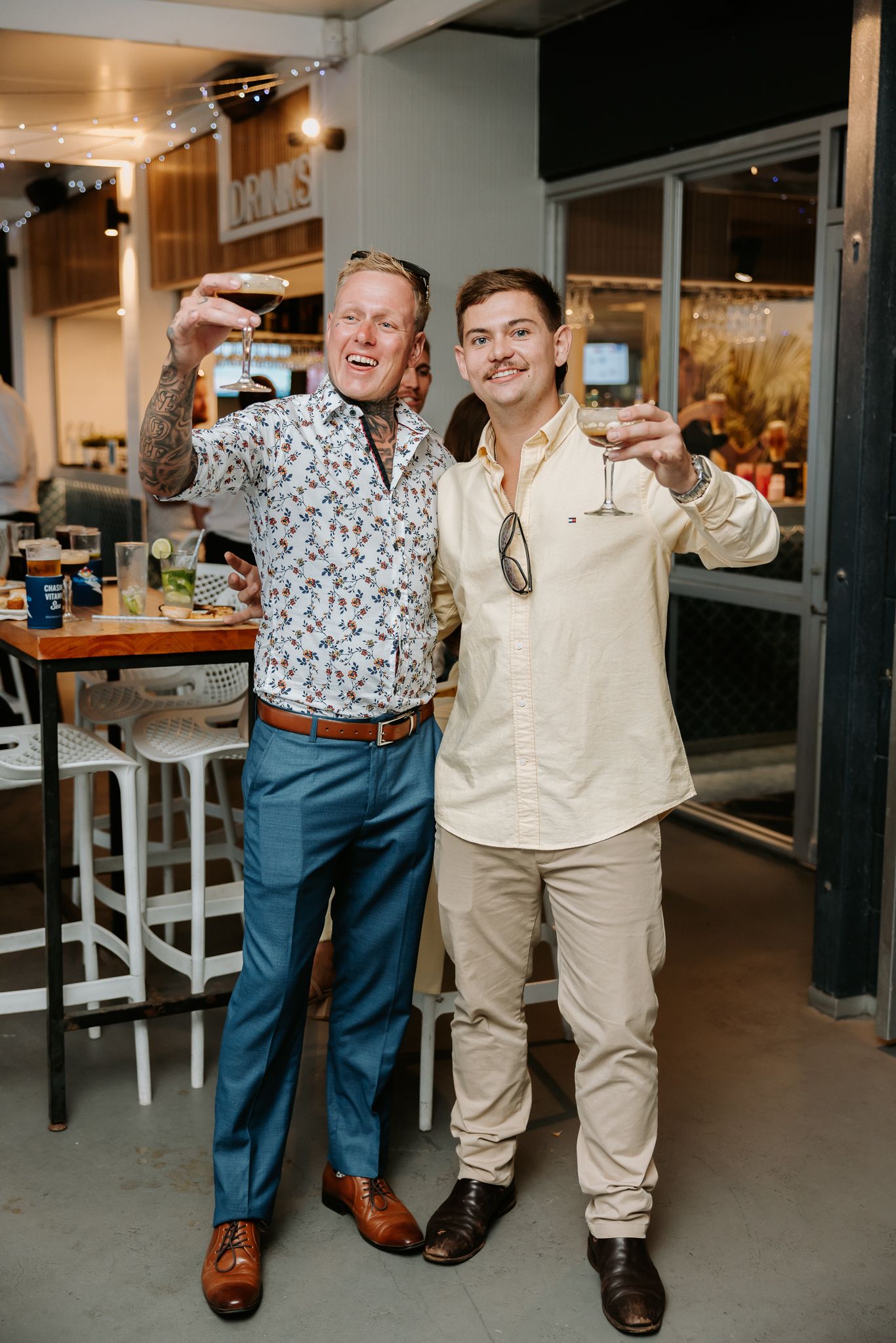 Two men raising cocktail glasses in a toast at a casual wedding reception bar area.