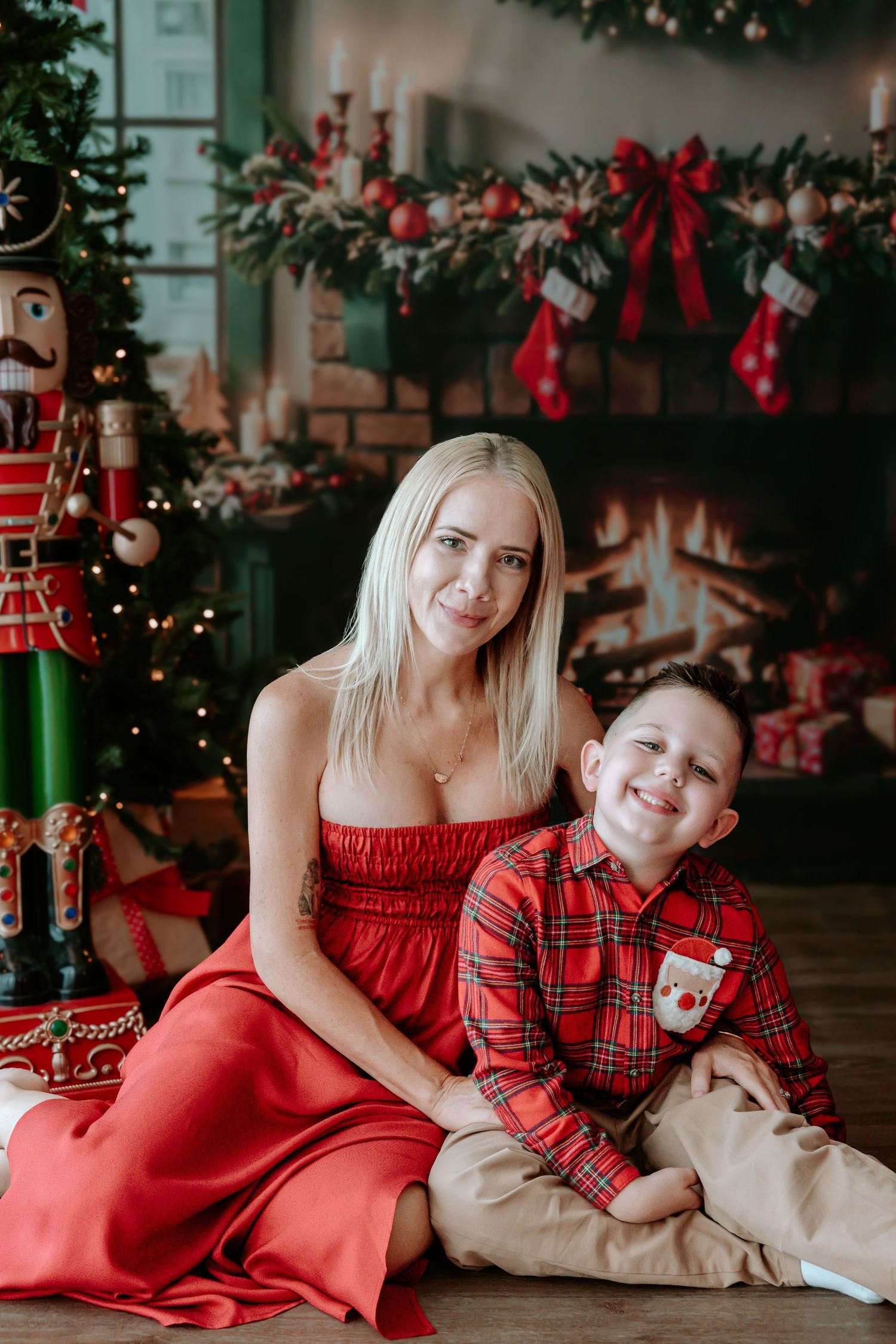 A smiling mother in a red dress poses with her young son in plaid shirt by a decorated Christmas fireplace.