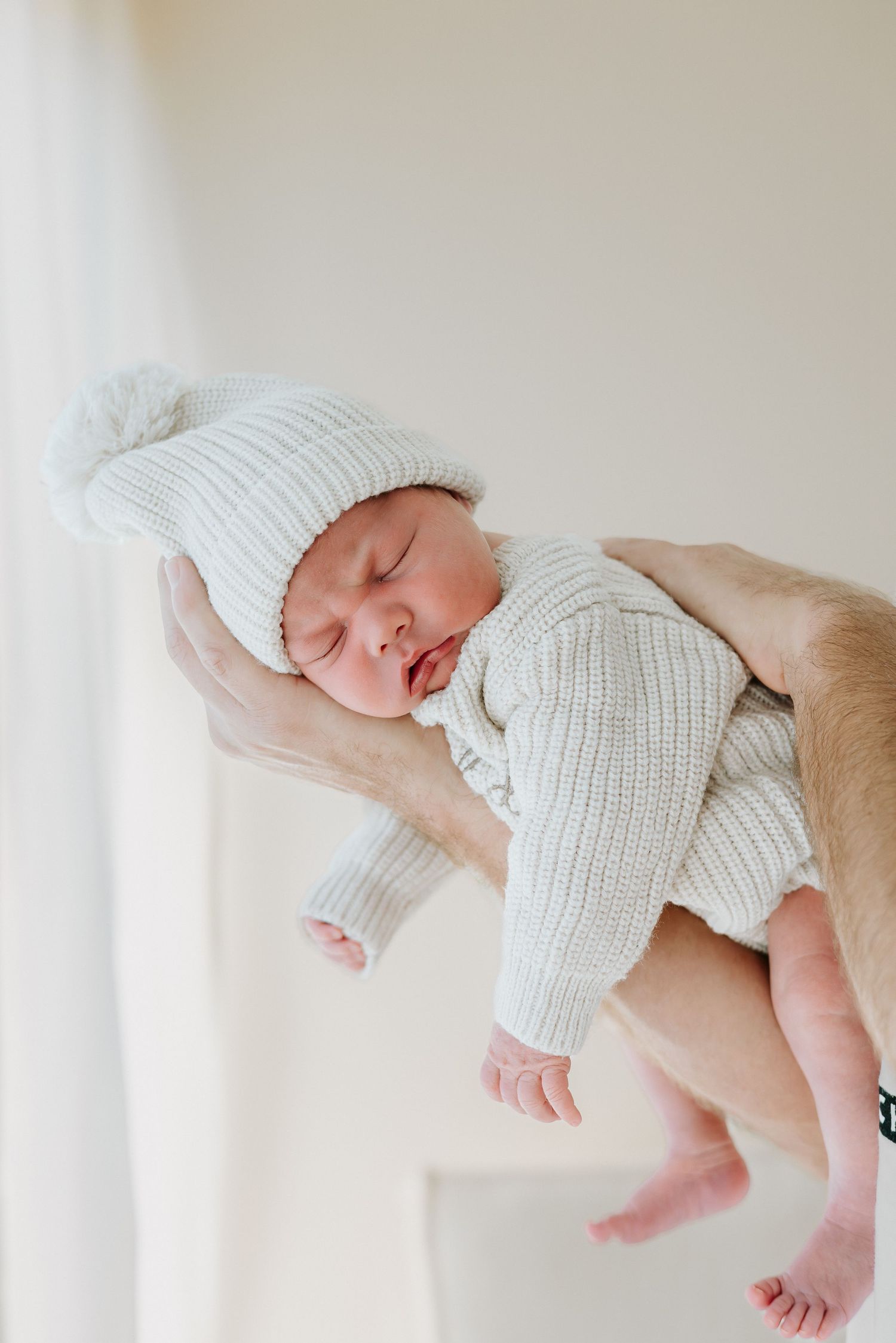 Newborn baby sleeping peacefully in white knit outfit and hat, cradled in adult hands against soft white background.