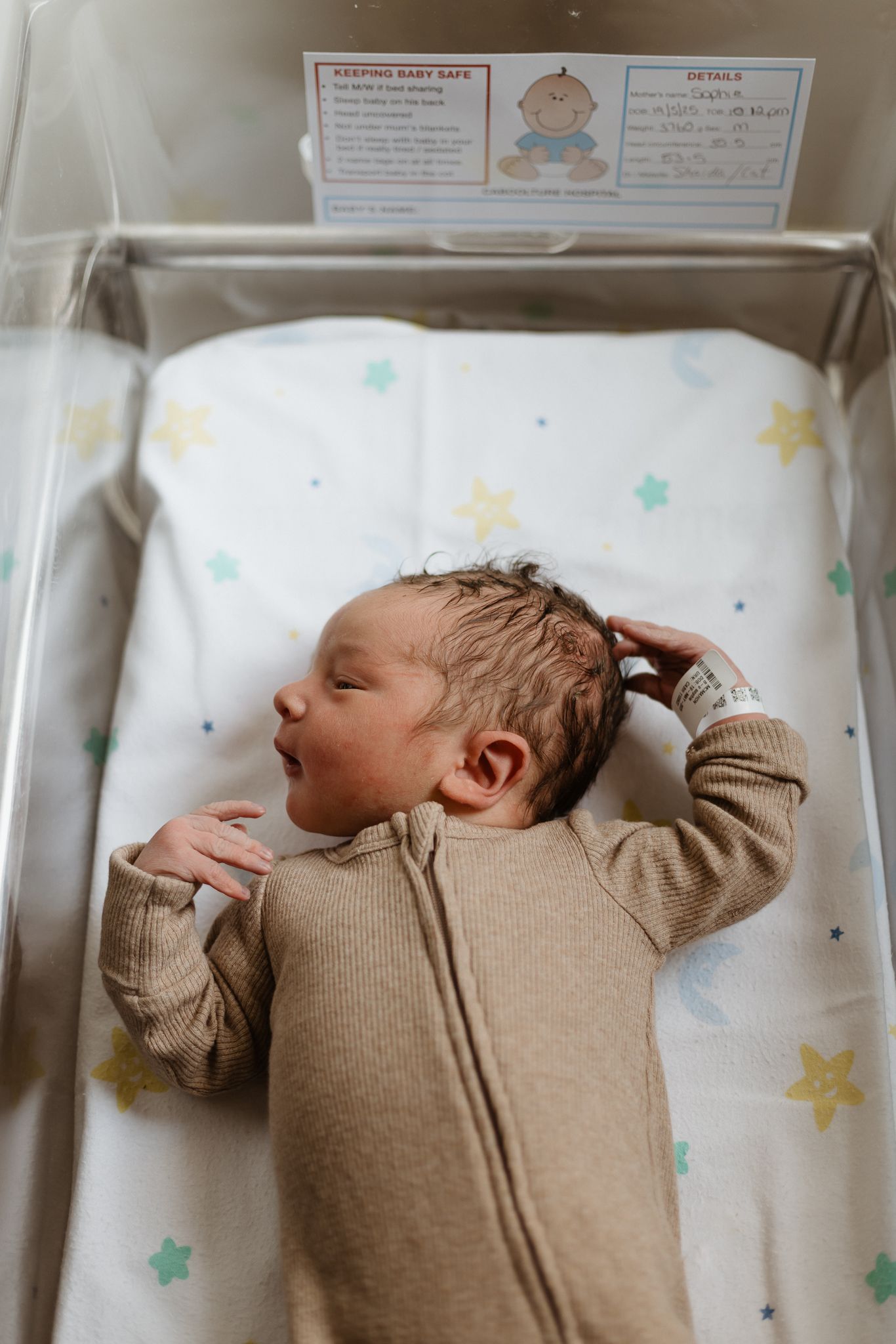 A newborn baby wearing a tan onesie lies on a changing pad decorated with pastel stars.