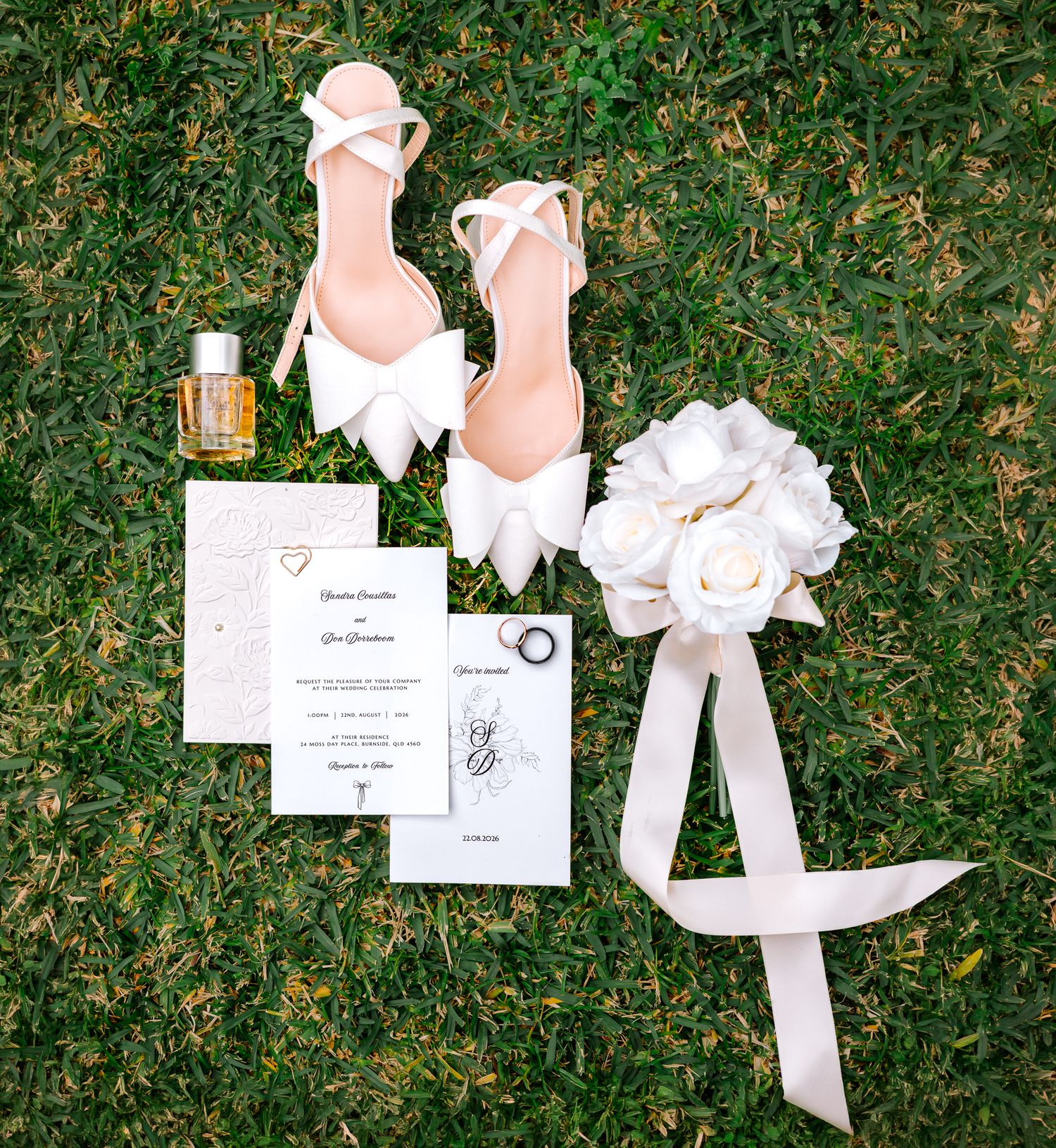 Elegant wedding flat lay featuring blush heels with bows, floral bouquet, invitations, and perfume on green grass.