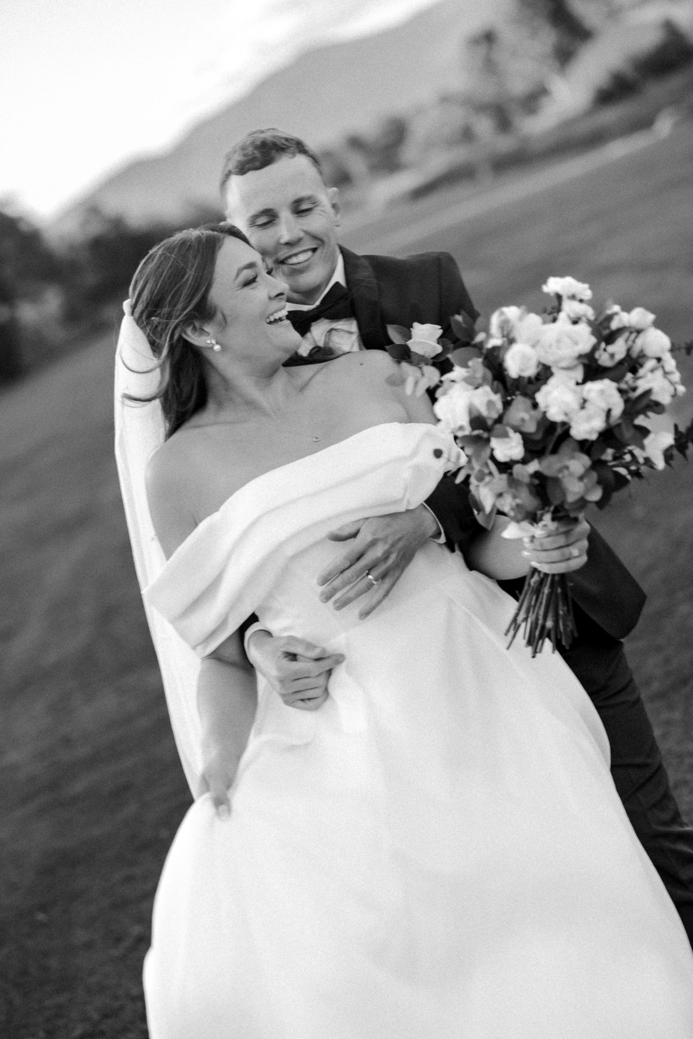 Black and white photo of a groom embracing his bride in a wedding dress holding a bouquet outdoors.