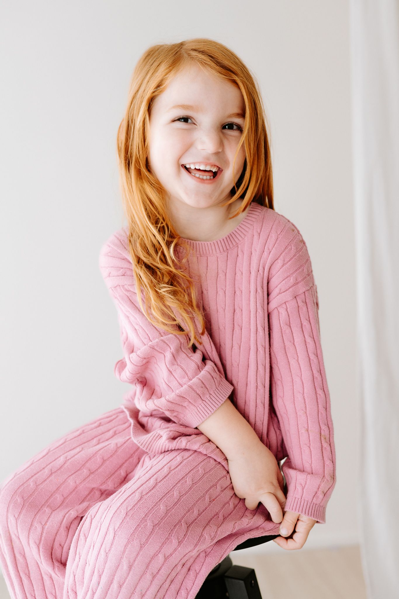 A cheerful young girl with long red hair wearing a pink cable-knit sweater set, smiling brightly while seated.