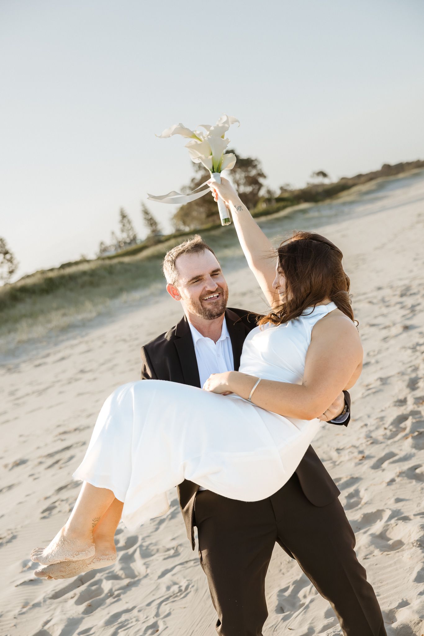 Groom carries bride in white dress on sandy beach, she holds bouquet aloft, both smiling joyfully.