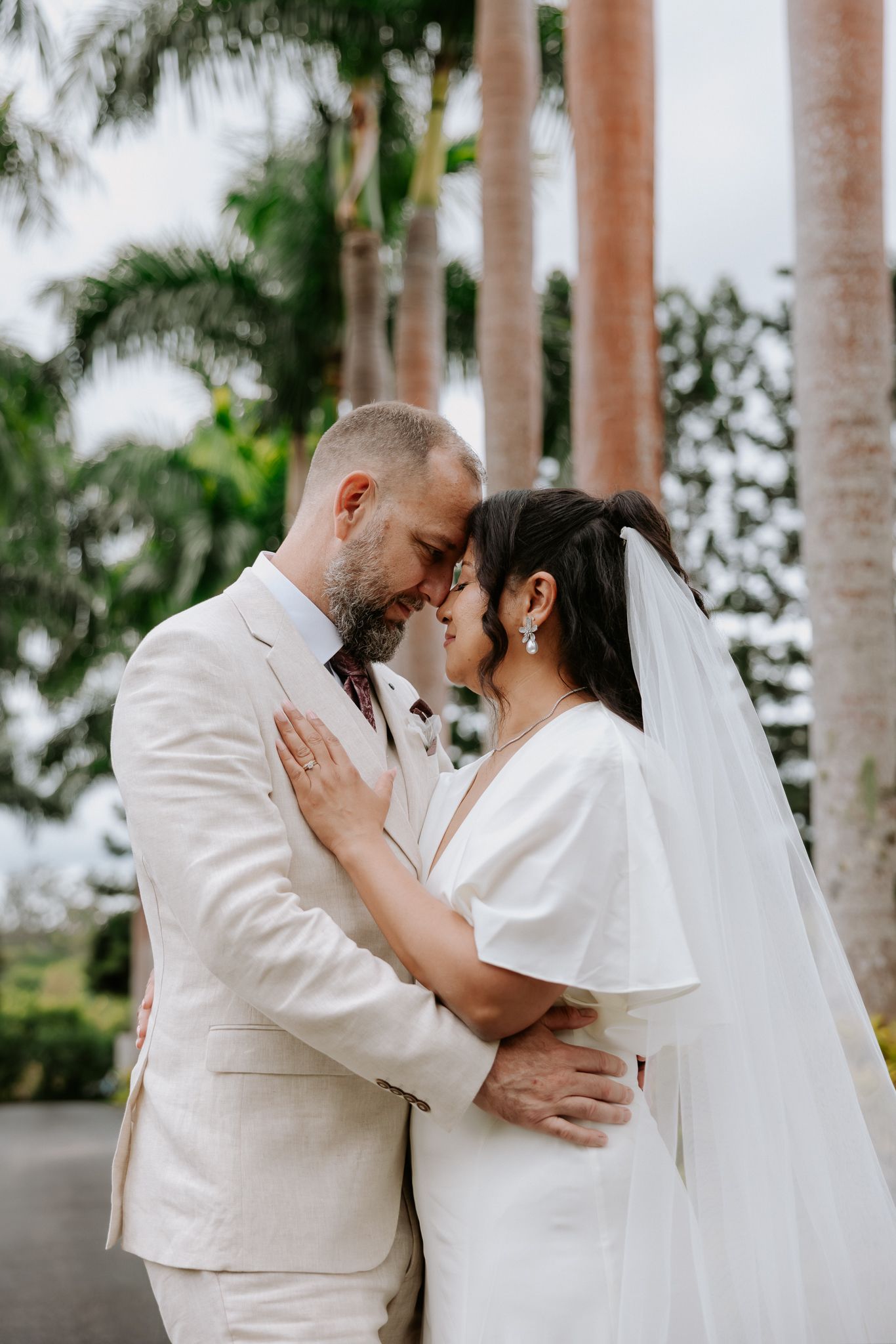 A couple in wedding attire share an intimate moment, foreheads touching, surrounded by tropical palm trees.
