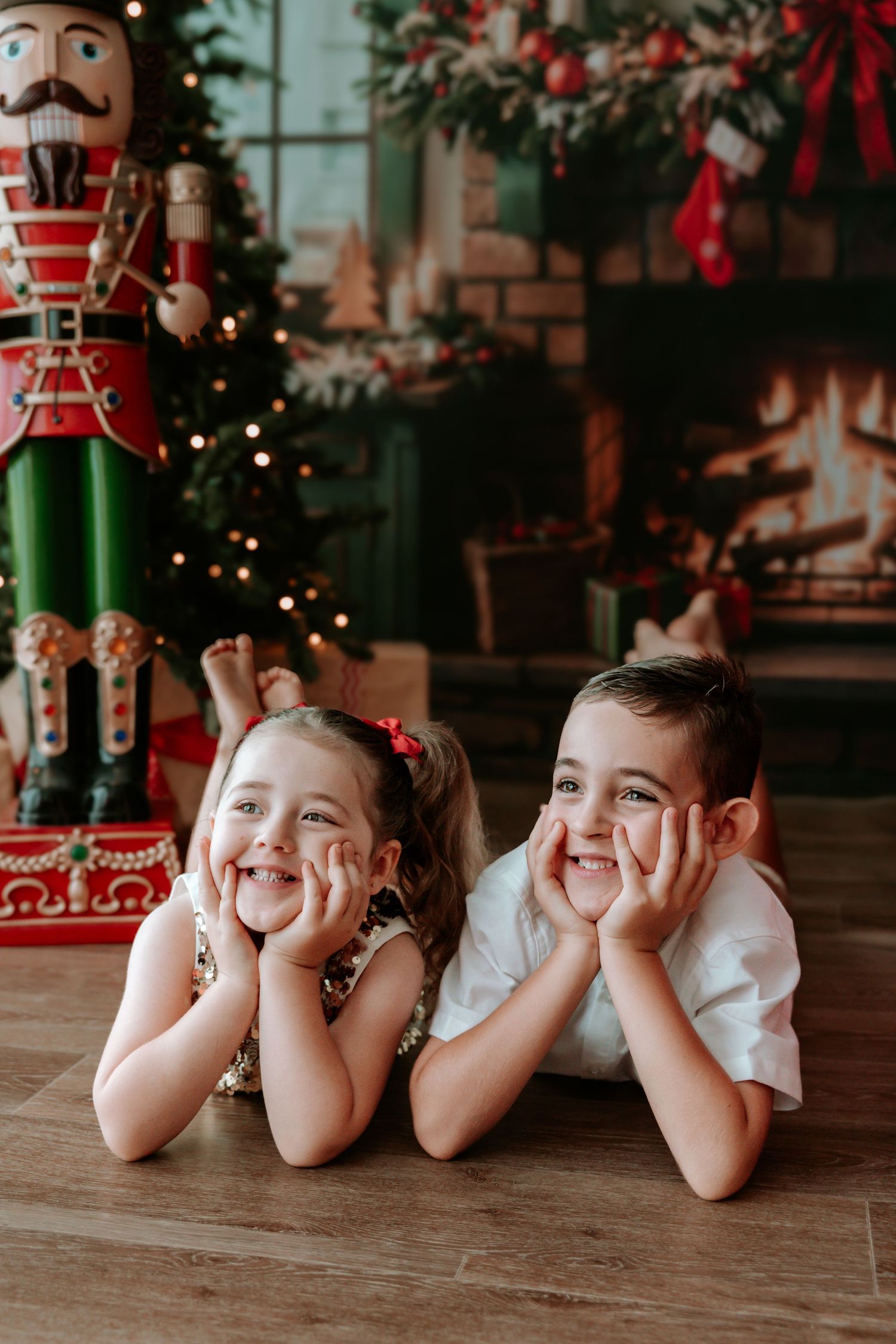 Two smiling children lying on floor in front of Christmas tree and nutcracker during holiday photo session.