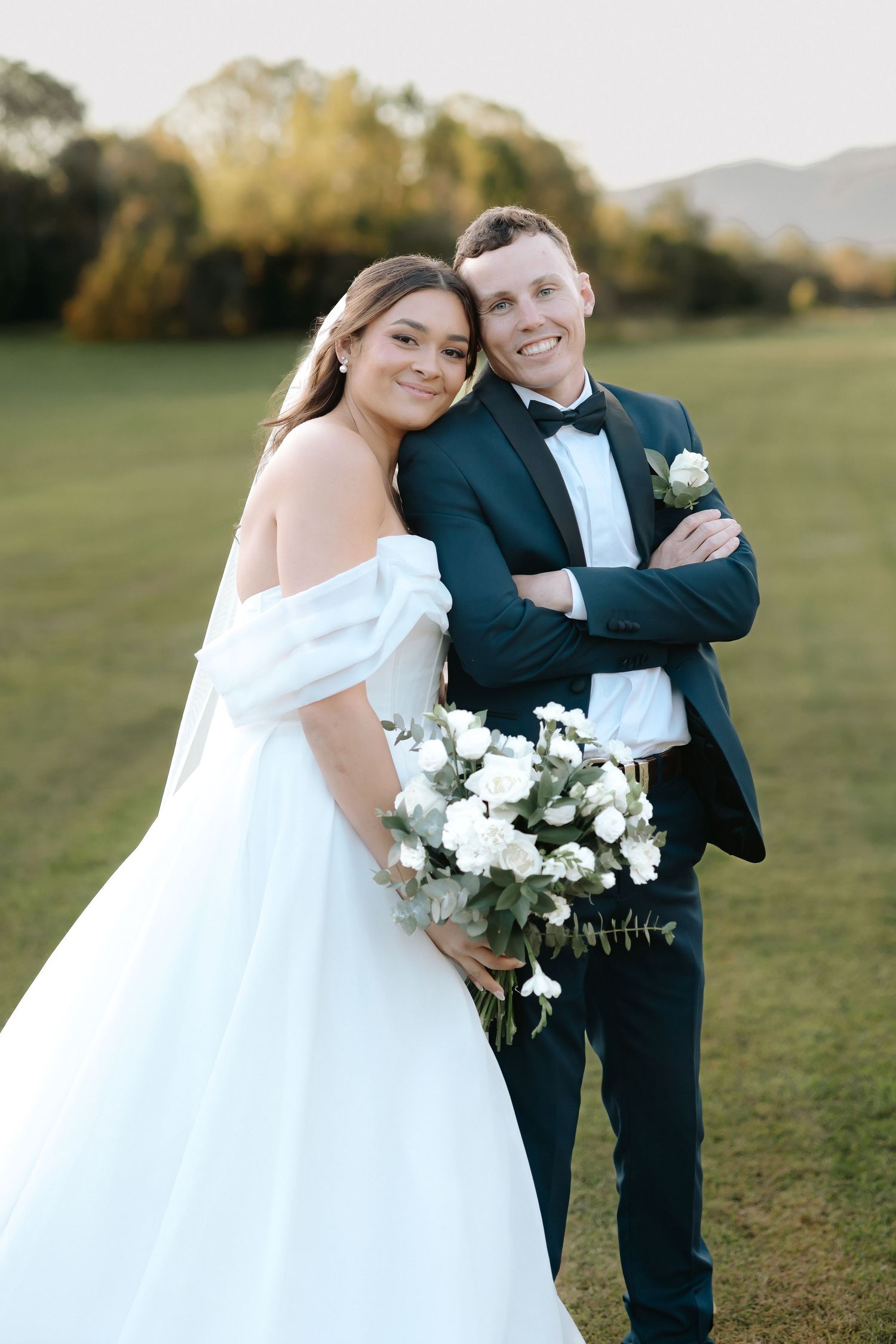 Bride in off-shoulder white gown holds bouquet leaning on groom in dark tuxedo outdoors at golden hour.