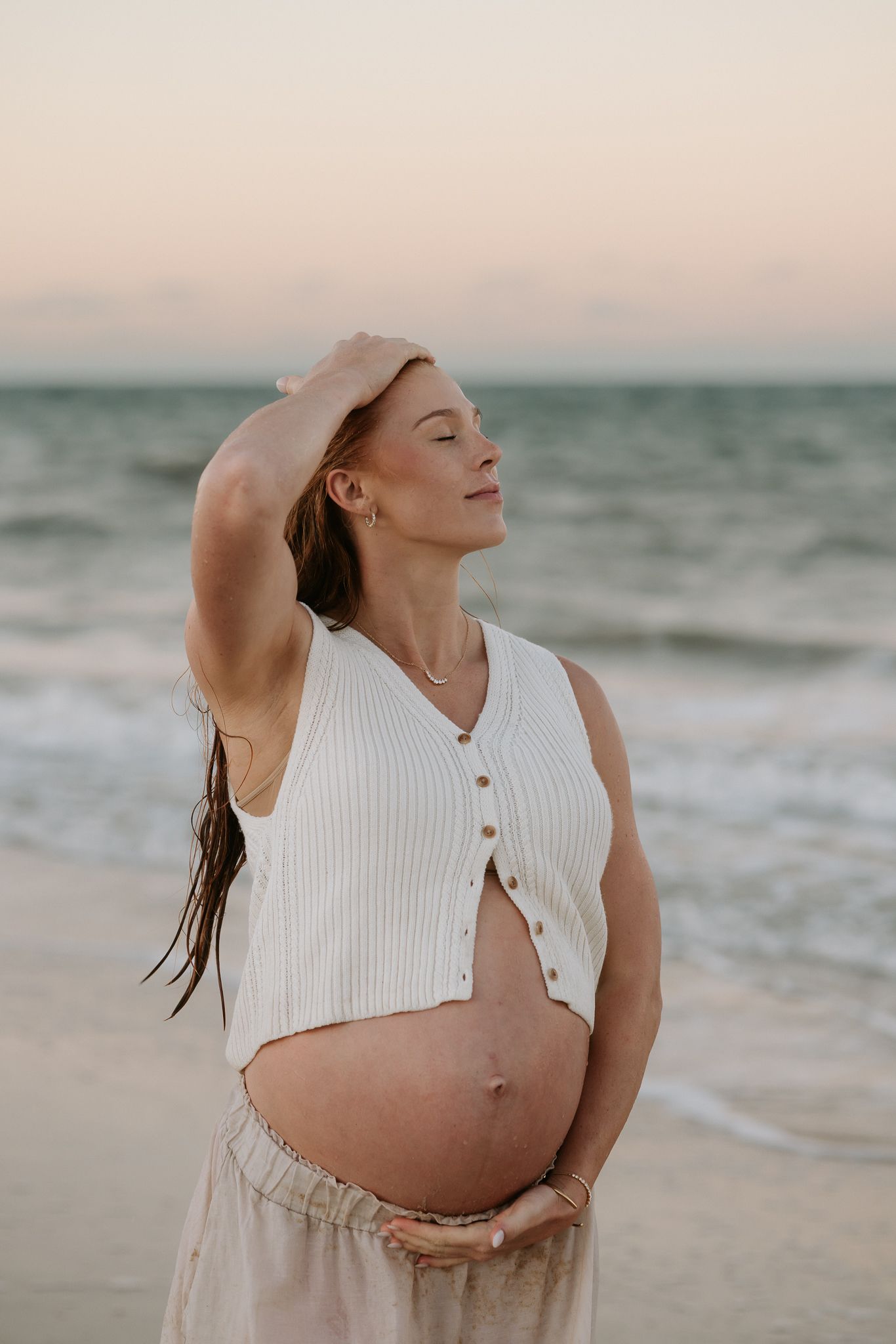 Pregnant woman in white crop top holds belly at beach during sunset, eyes closed, hand raised to head.