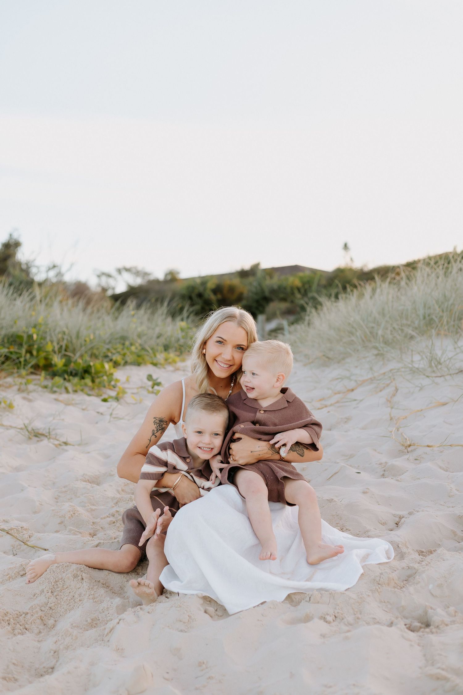 A smiling blonde mother sits on sandy beach dunes hugging two young toddler boys dressed in brown outfits.