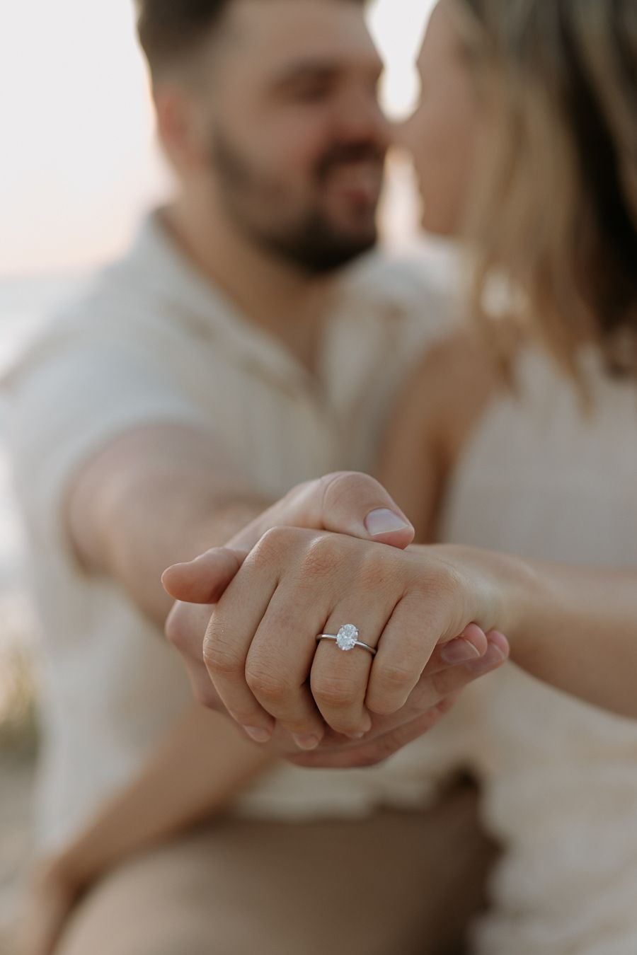 Couple holding hands showing oval diamond engagement ring in foreground, romantic beach proposal moment.
