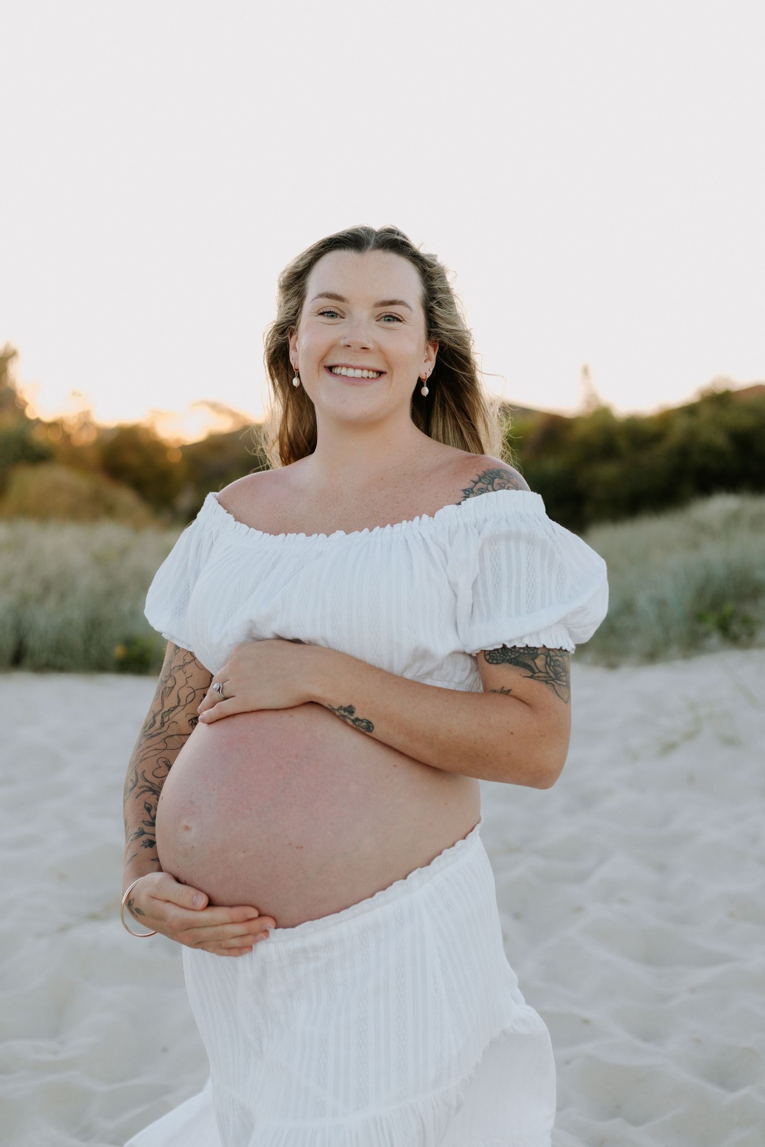 Expectant mother in white off-shoulder dress poses on sandy beach at sunset showing her baby bump.