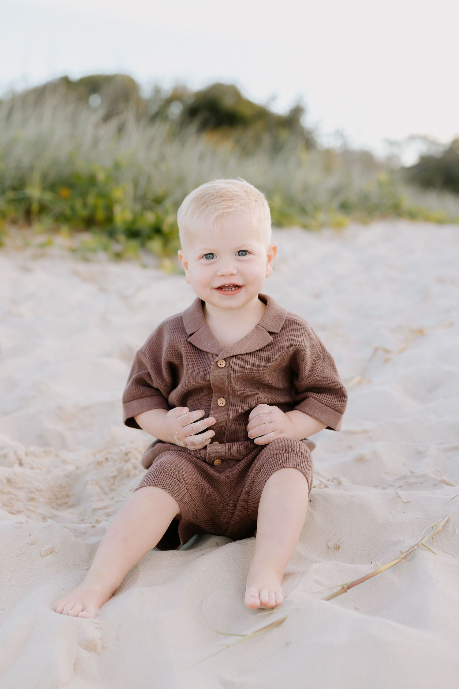 Smiling toddler in brown romper sitting on sandy beach with dunes and greenery in background.