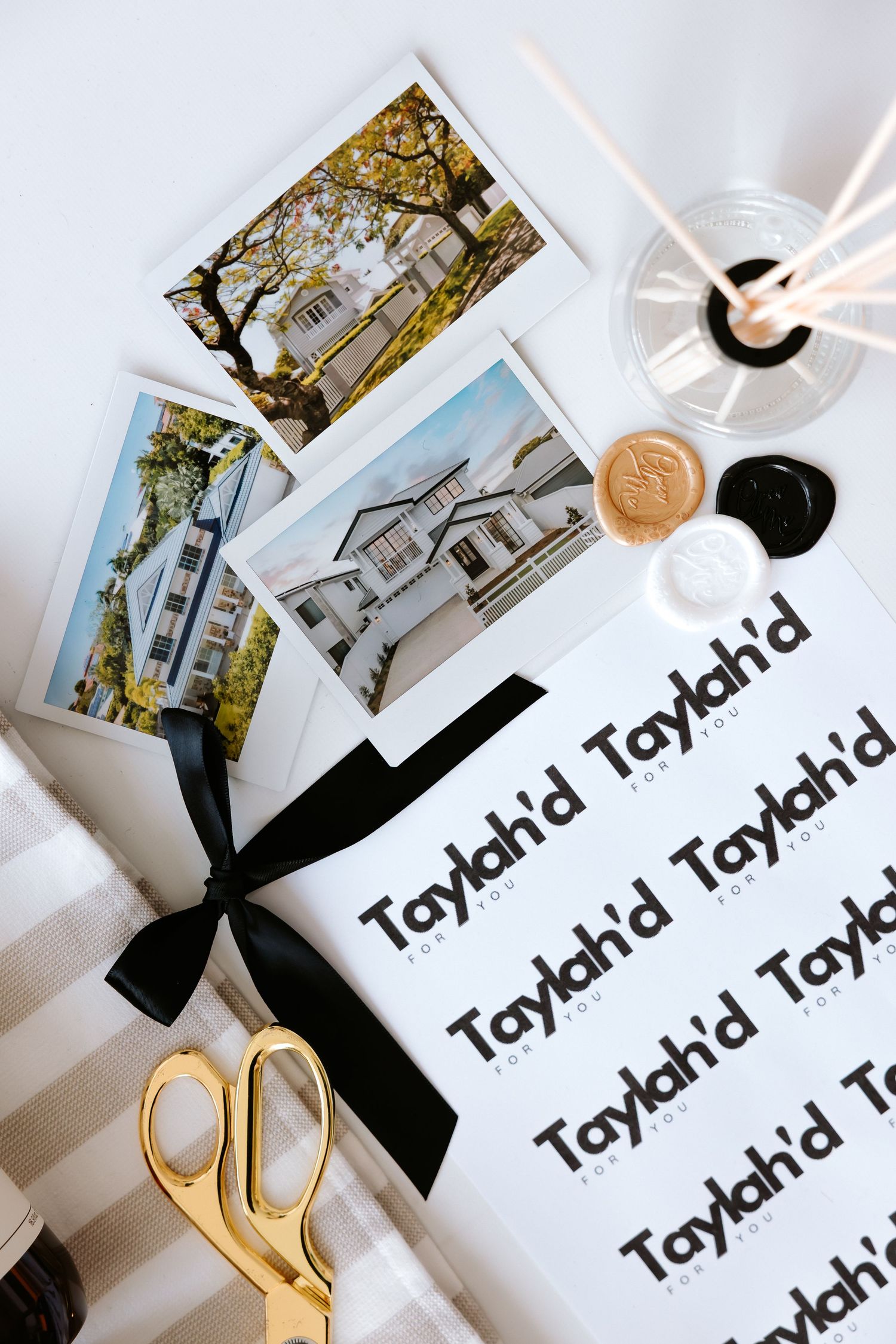 Flat lay of Taylah'd branding stationery with property photos, black ribbon, gold scissors, wax seals, and a champagne glass.