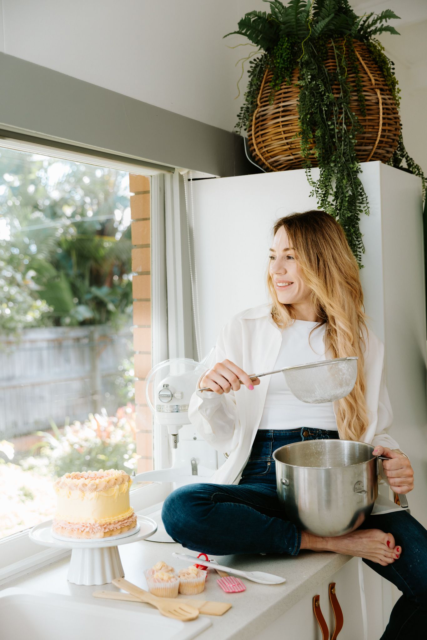 Smiling woman with long blonde hair sitting on kitchen counter with mixing bowl, cake, and cupcakes near bright window.